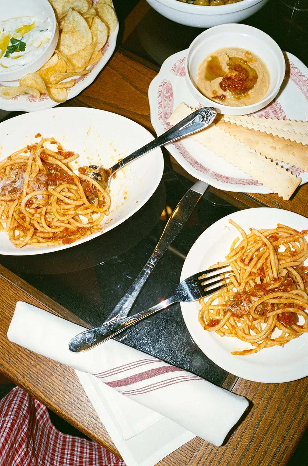 Two plates of spaghetti with tomato sauce, a bowl of potato chips with a dip, and a bowl of curry with flatbread on a wooden table.