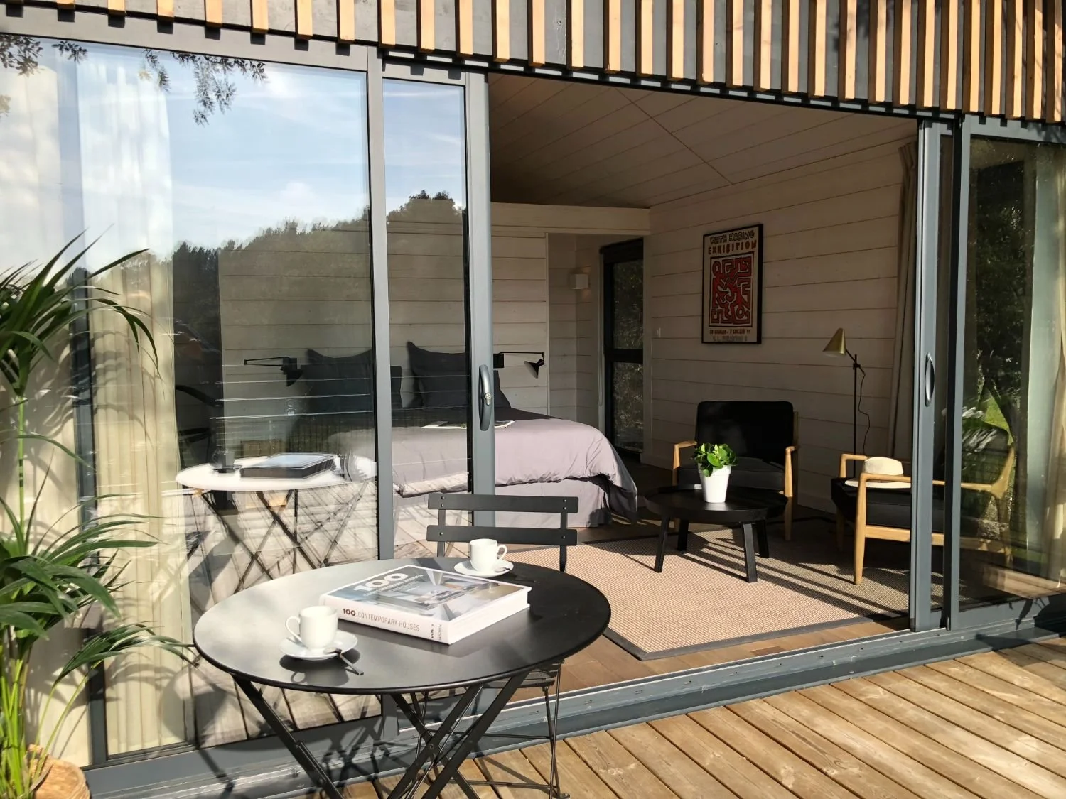 Un intérieur moderne avec un lit, un petit meuble rond avec deux tasses, une plante en pot, une chaise, un fauteuil, un lampadaire et une affiche sur le mur, visible à travers une porte-fenêtre.