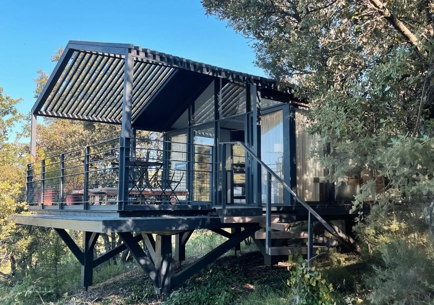 Une tiny house en bois noire avec une terrasse entourée de végétation et de arbres, des escaliers menant à la porte d'entrée, et une toiture avec des lattes en bois.