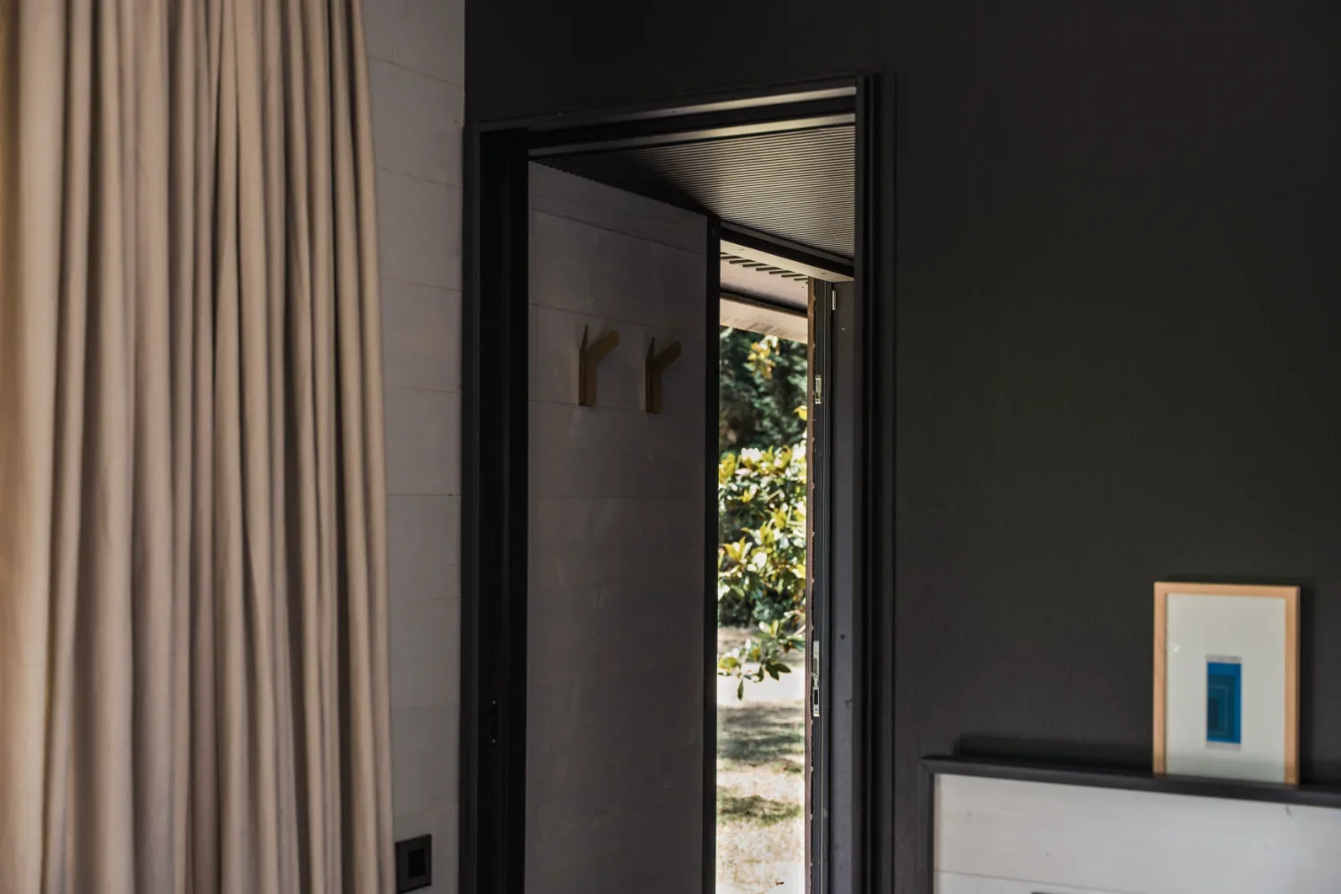 Une porte entrouverte laissant entrevoir un extérieur avec des arbres et des plantes, dans une pièce moderne avec des murs sombres et des décorations minimalistes.