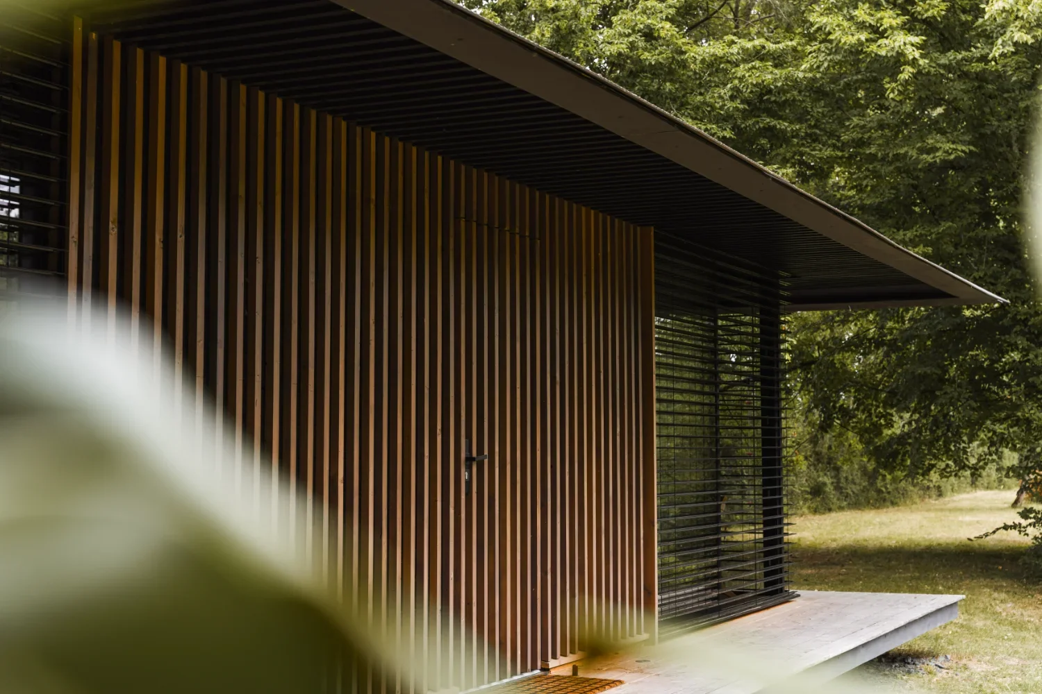 Façade moderne d'un Ecolodge TESHIMA avec des volets en lattes et une avancée en béton, entourée d'arbres.