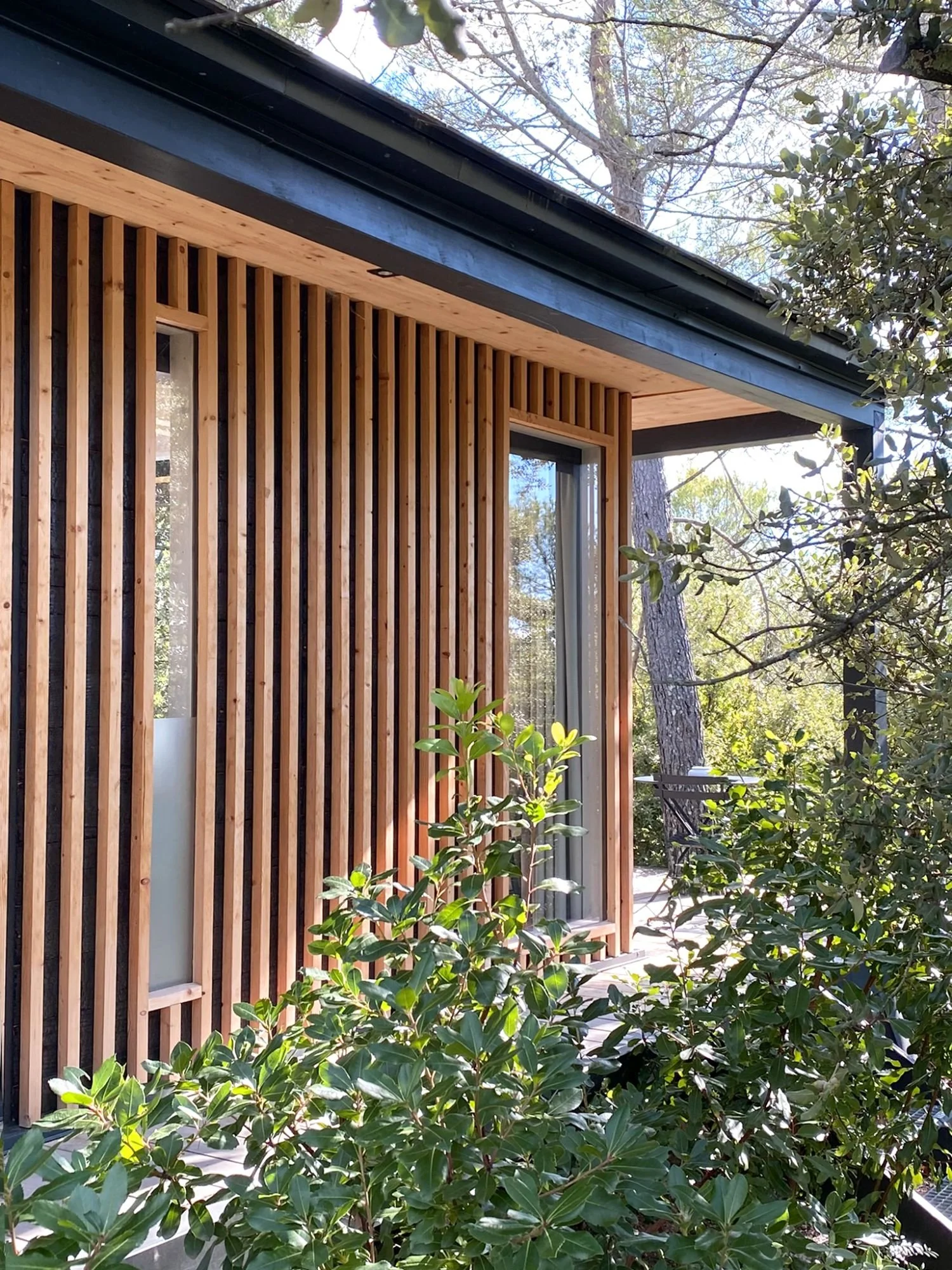 Façade en bois d'un Ecolodge KOBE avec des murs en lattes verticales, grande fenêtre et terrasse entourée de verdure et d'arbres.