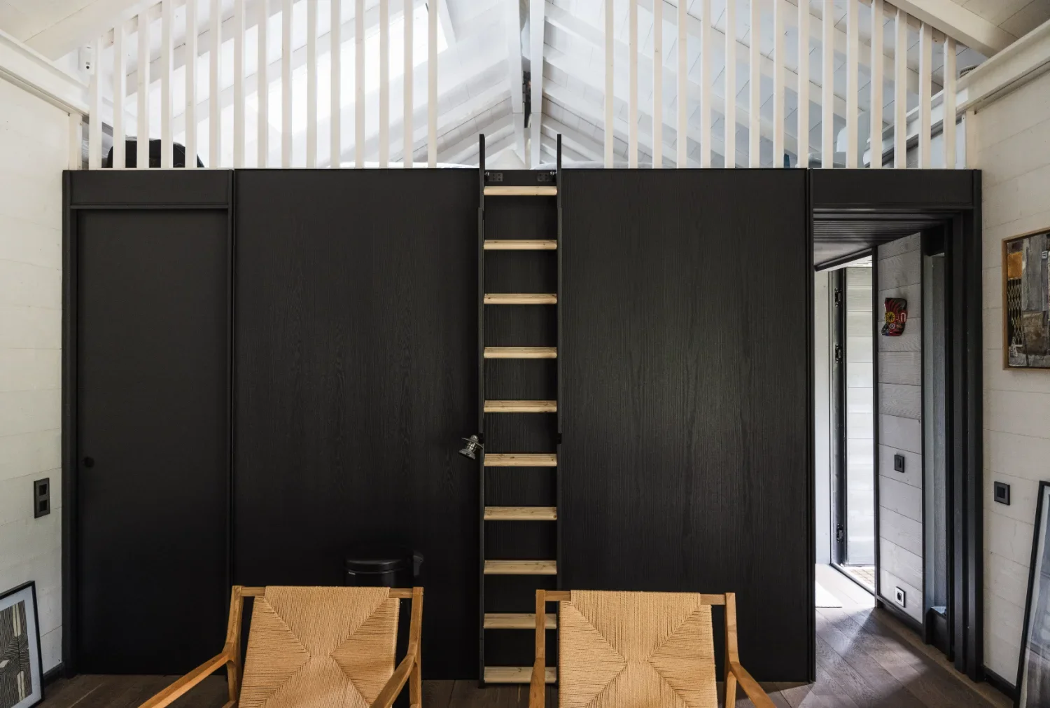Intérieur d'un Ecolodge TESHIMA avec un mur noir, une échelle en bois menant à un espace supérieur, un coin salon avec deux fauteuils en bois et une porte vitrée à côté, dans un intérieur moderne.
