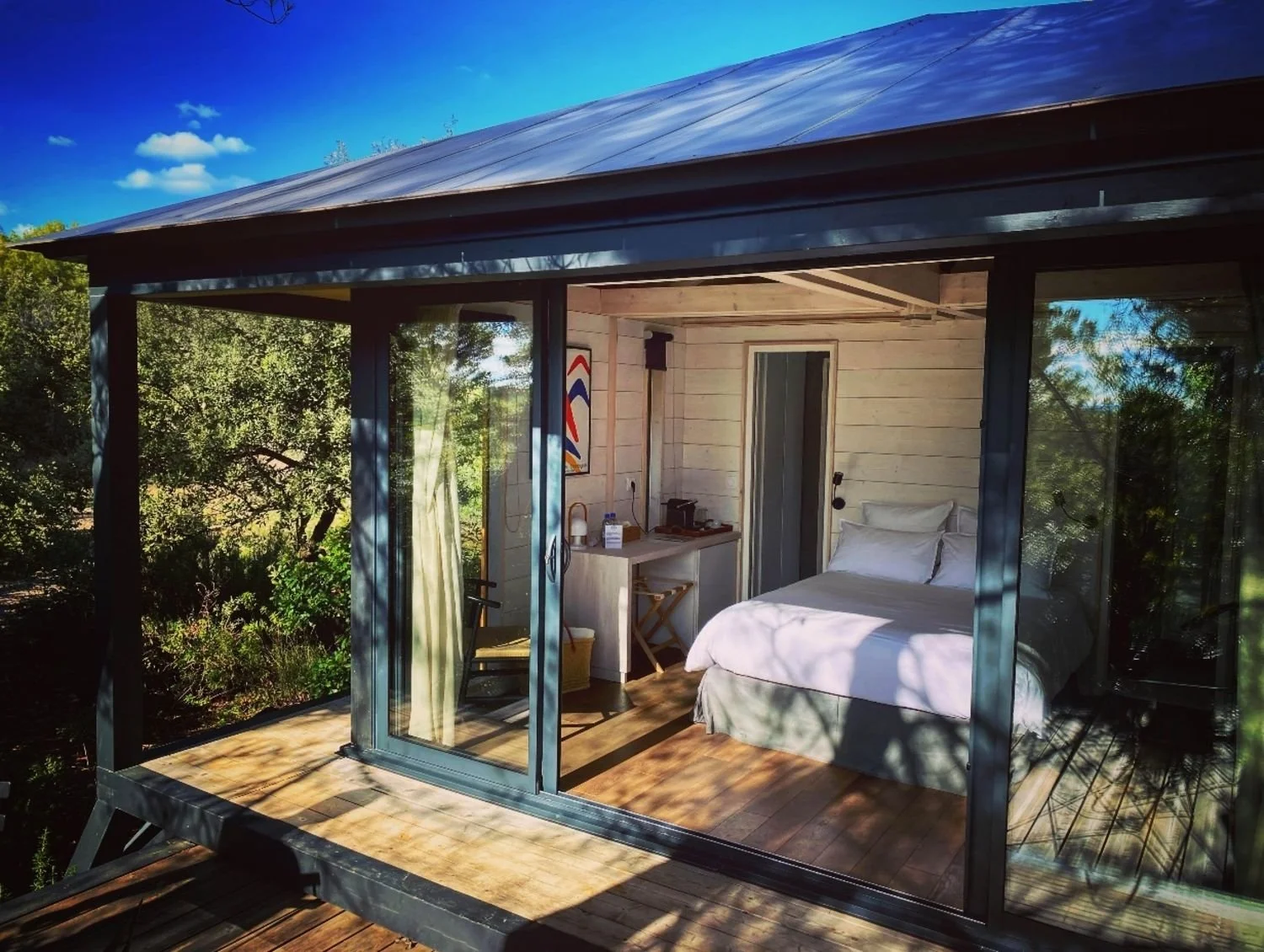Ecolodge KOBE avec un lit blanc et un petit coin bureau, située dans un cadre naturel avec des arbres et un ciel bleu.