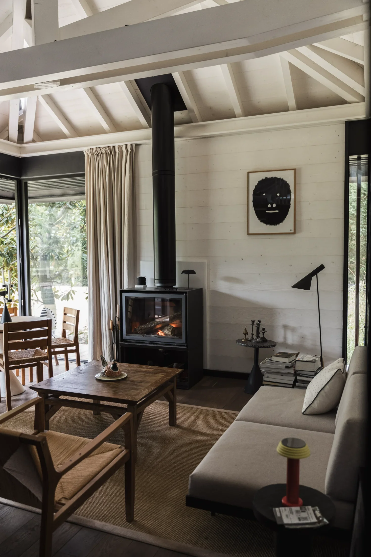 Salon intérieur d'un Ecolodge TESHIMA avec cheminée à bois, canapé beige, table en bois, livres empilés à côté du canapé, lampe noire, tableau noir et blanc sur le mur, rideaux beiges, grande fenêtre laissant entrer de la lumière naturelle.