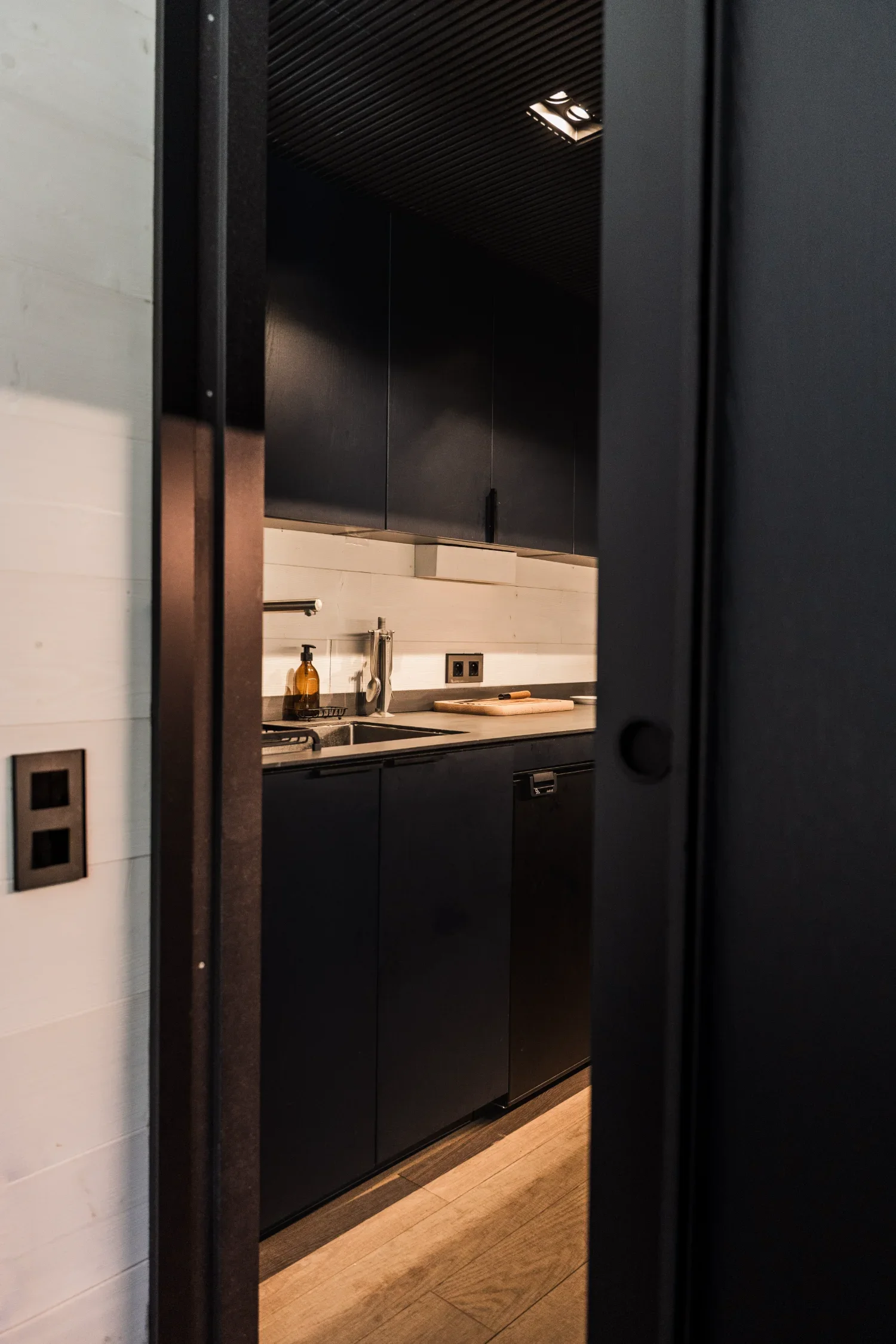 Une cuisine moderne avec des armoires noires, un évier, un dosseret blanc, une bouteille en verre, un liquide vaisselle, une planche à découper, une prise électrique, probablement vue depuis une porte entrouverte.