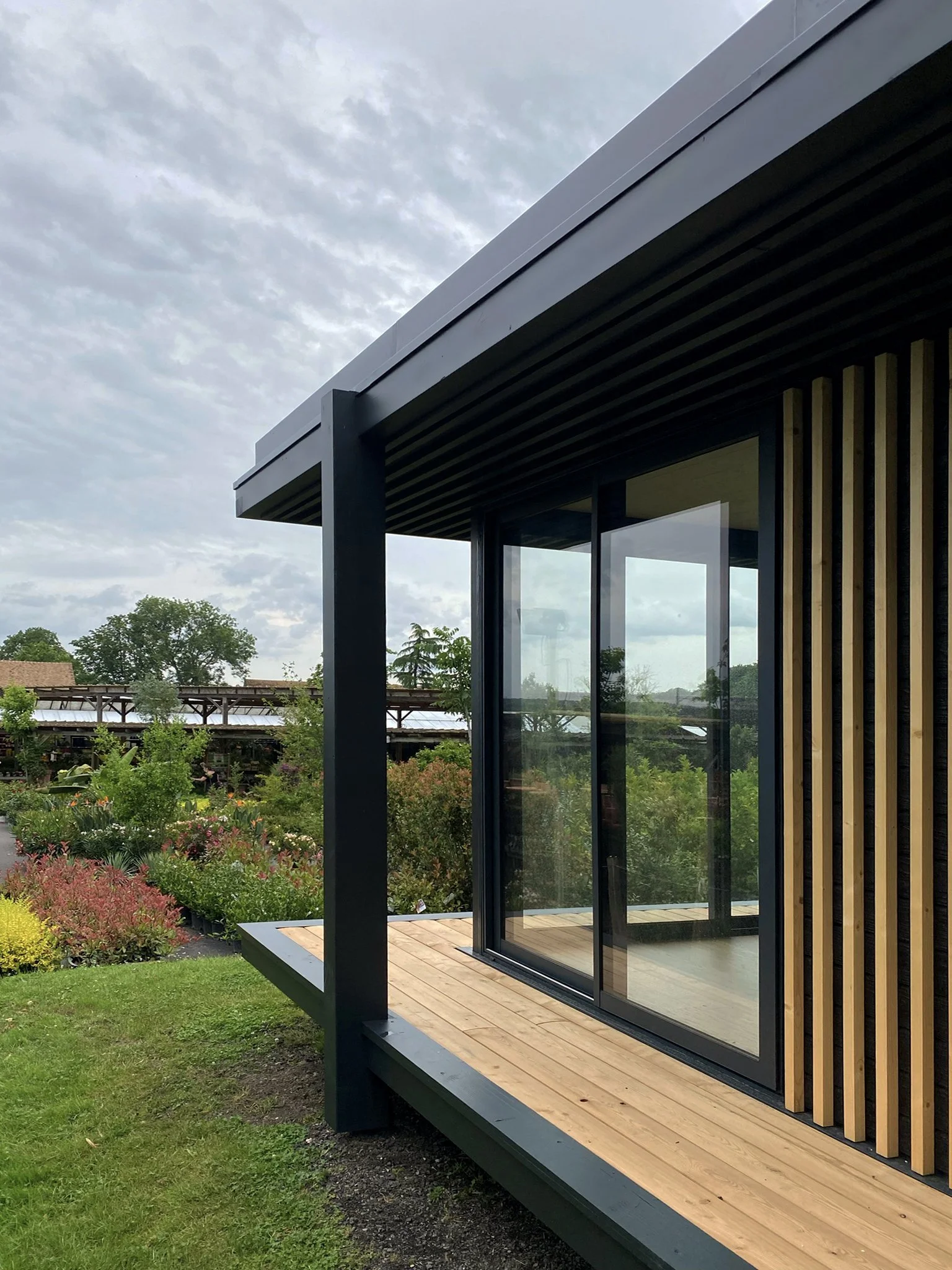 Vue d'un Ecolodge MEGYMA avec une terrasse en bois, murs en verre et un design en lames de bois verticales, entourée de verdure et de jardins.