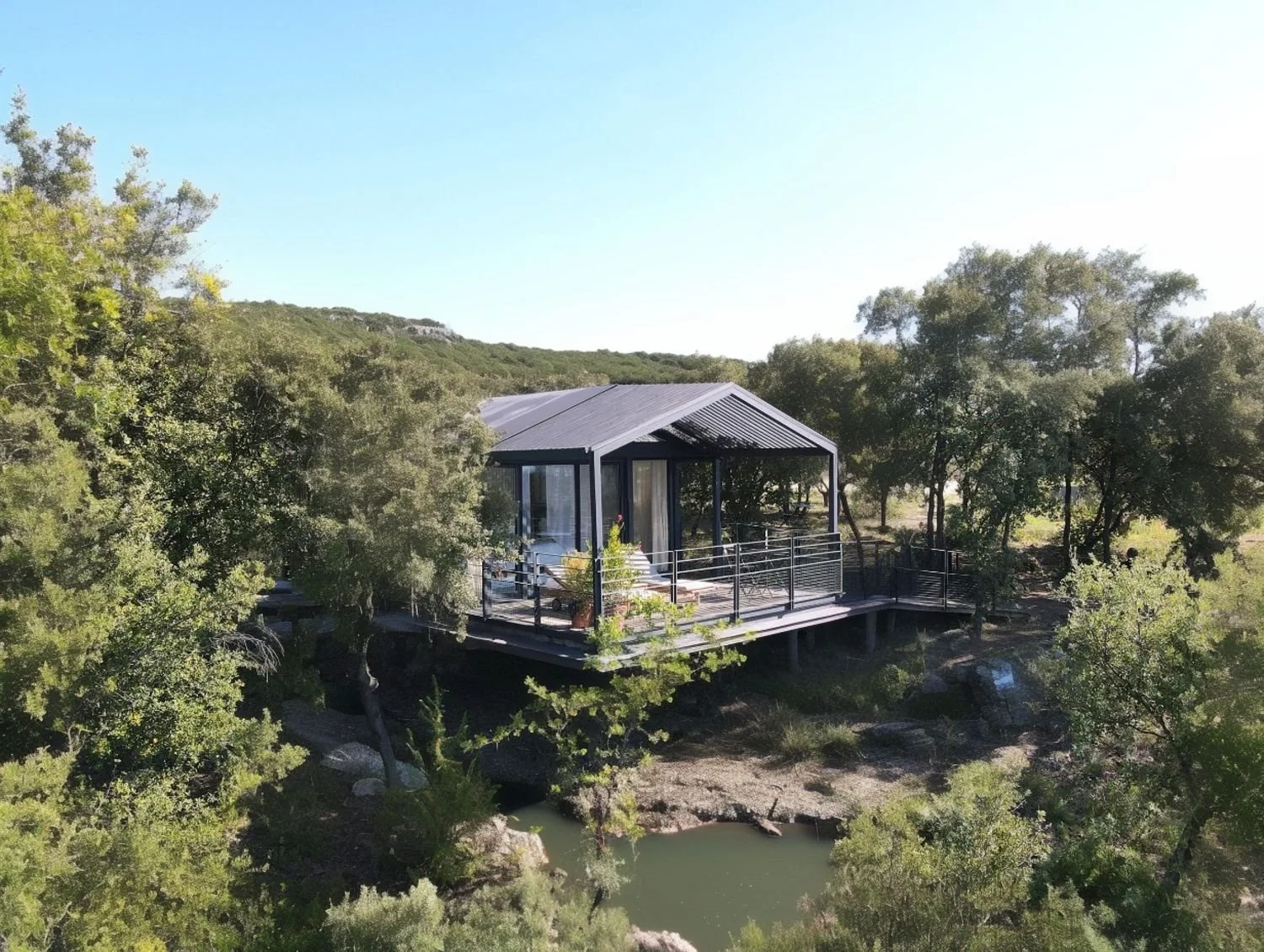 Cabane moderne en métal noir, perchée au-dessus d'une rivière, entourée d'arbres verts, sous un ciel bleu clair.