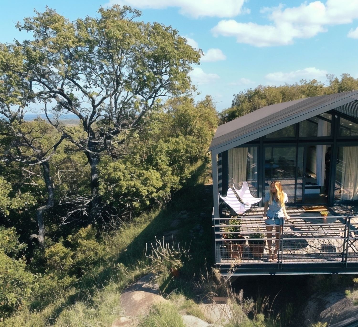 Une maison moderne en verre à été construite sur une falaise en hauteur avec une vue sur la nature et des arbres verts. Sur la terrasse, une femme blonde en jean et une personne en pantalon noir parlent, il y a des plantes en pot et une chaise suspen