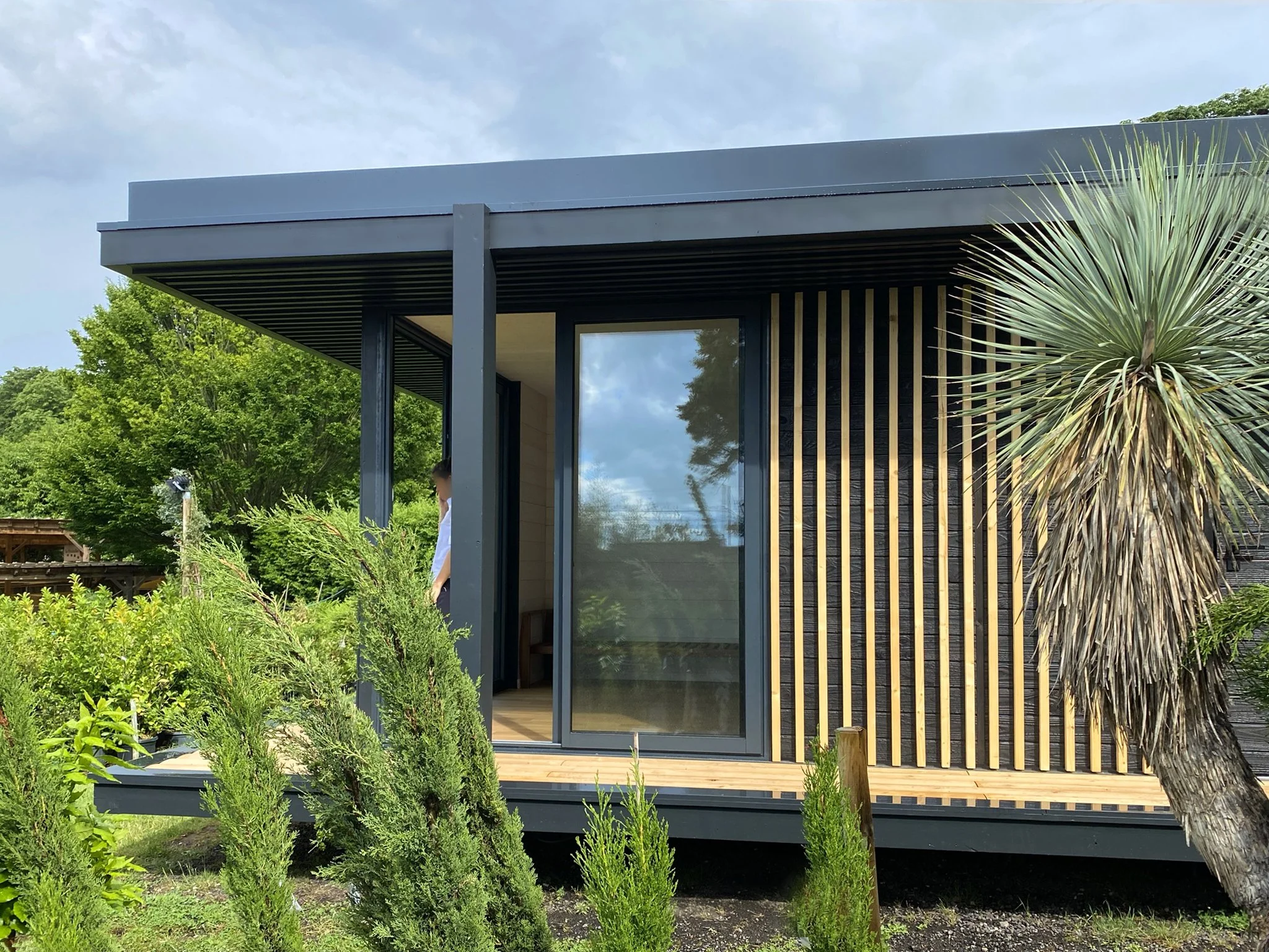 Ecolodge MEGYMA avec une grande fenêtre en verre, entourée de végétation, dont un palmier. Le ciel est partiellement nuageux.