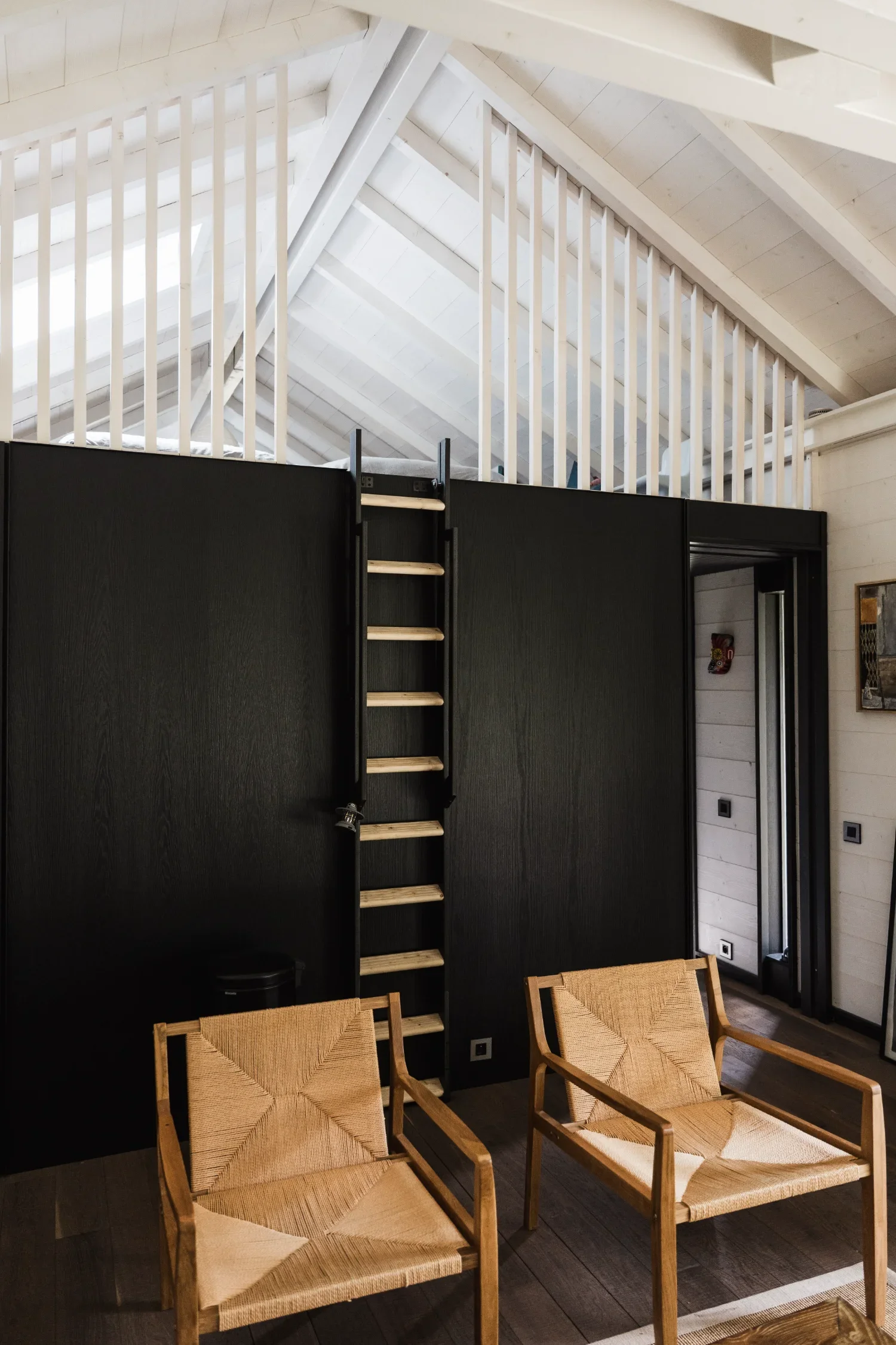 Deux fauteuils en bois avec coussins en corde tissée devant un mur noir dans un intérieur lumineux avec plafond en bois blanc et escalier en bois menant à un espace en mezzanine d'un Ecolodge TESHIMA