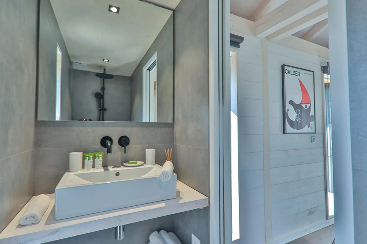 Salle de bain moderne avec un lavabo blanc, miroir, produits de toilette, serviettes et vue sur une douche avec tête de douche noire dans un environnement épuré en gris et blanc.