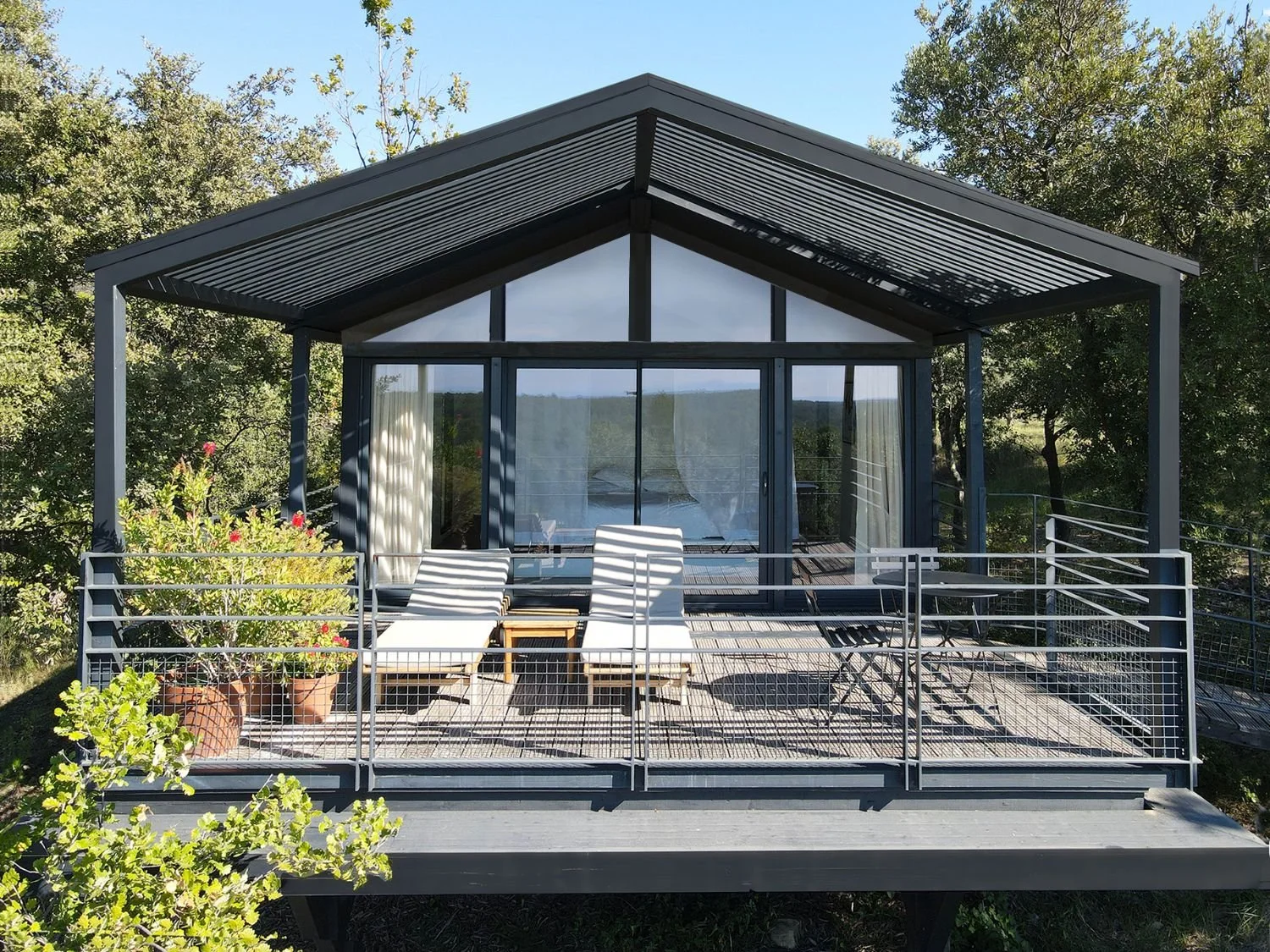 Une terrasse en bois avec un balcon entouré d'une barrière en métal, donnant sur un environnement verdoyant avec des arbres, une maison en verre avec un toit en pente et des rideaux blancs à l'intérieur.