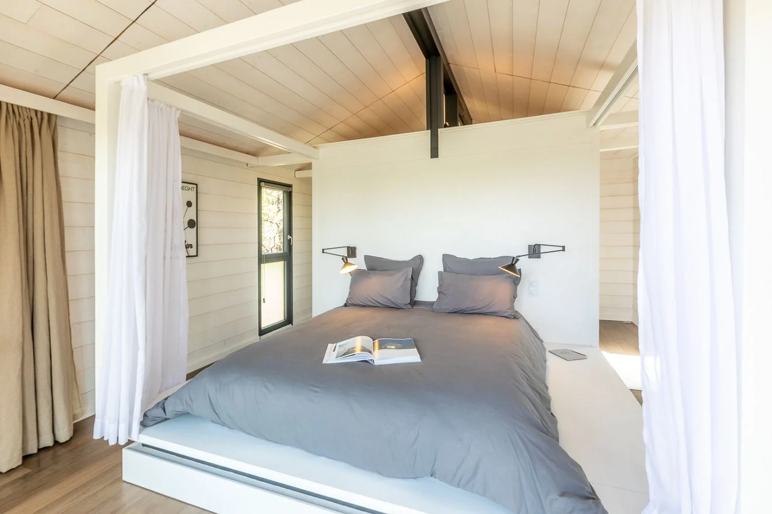 chambre moderne avec lit double, draps gris, table de nuit, lampes murales, fenêtres, murs en bois clair, porte-fenêtre, livres sur le lit, rideaux blancs et beige, plafond en bois