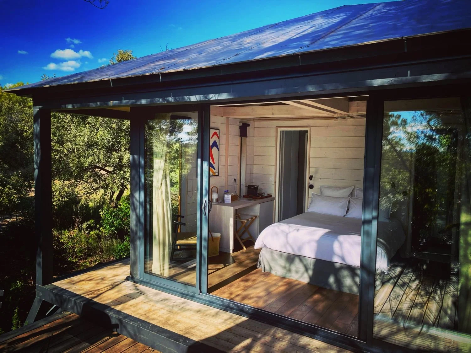 Ecolodge KOBE avec un mur en verre, un lit blanc, une petite table, et une vue sur la nature environnante, sous un ciel bleu avec quelques nuages.
