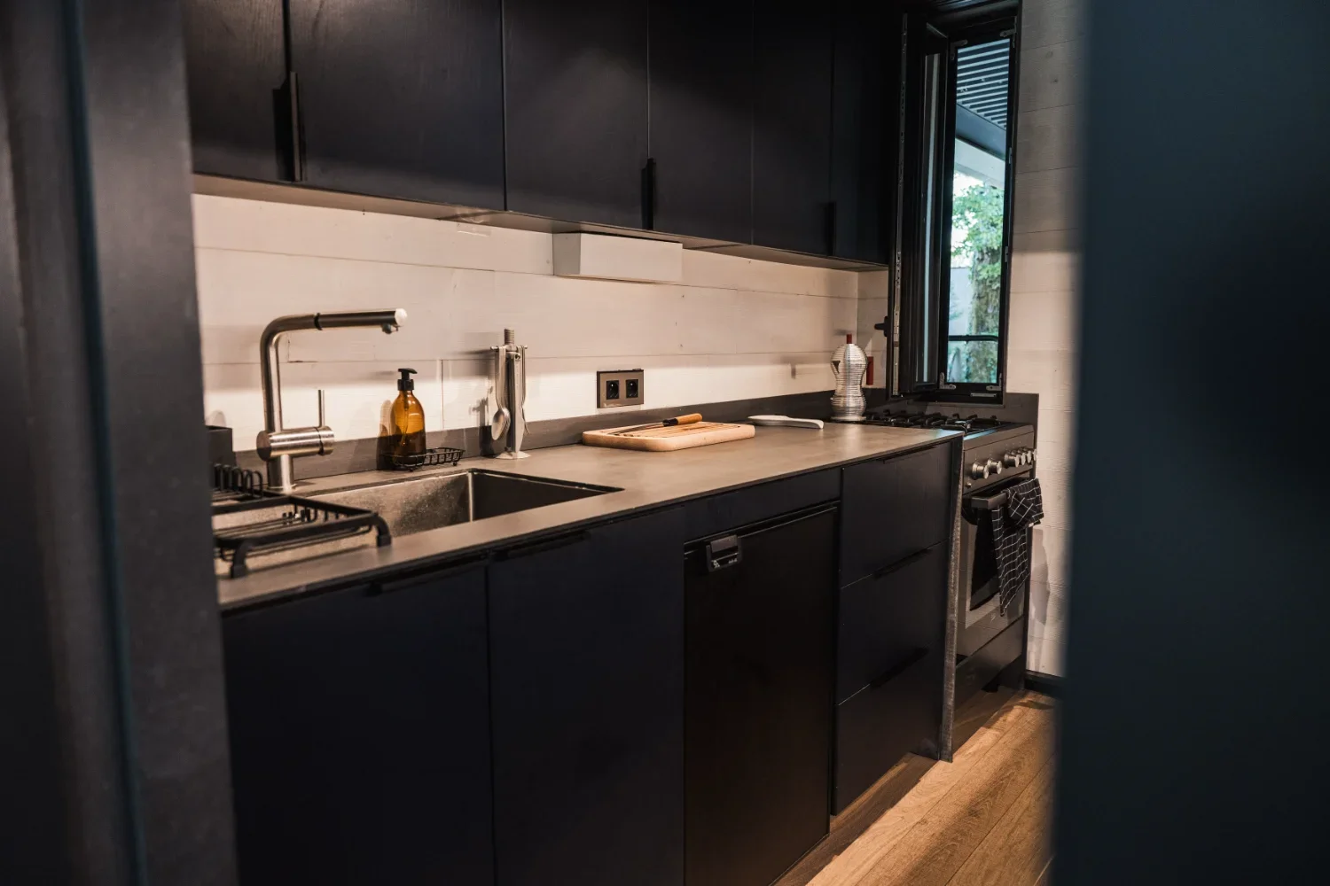 Cuisine moderne avec meubles noirs, évier, plan de travail, une bouteille d'eau, une planche à découper, et une fenêtre laissant entrer de la lumière naturelle.