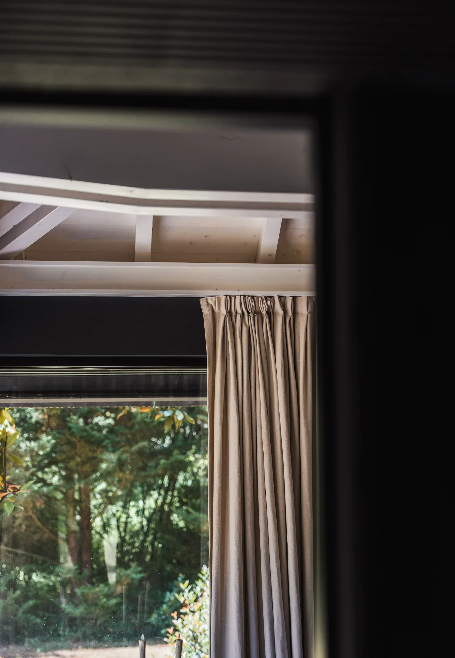 Vue depuis l'intérieur d'un Ecolodge TESHIMA, vue partielle d'une fenêtre avec rideau beige et vue sur un jardin avec des arbres.