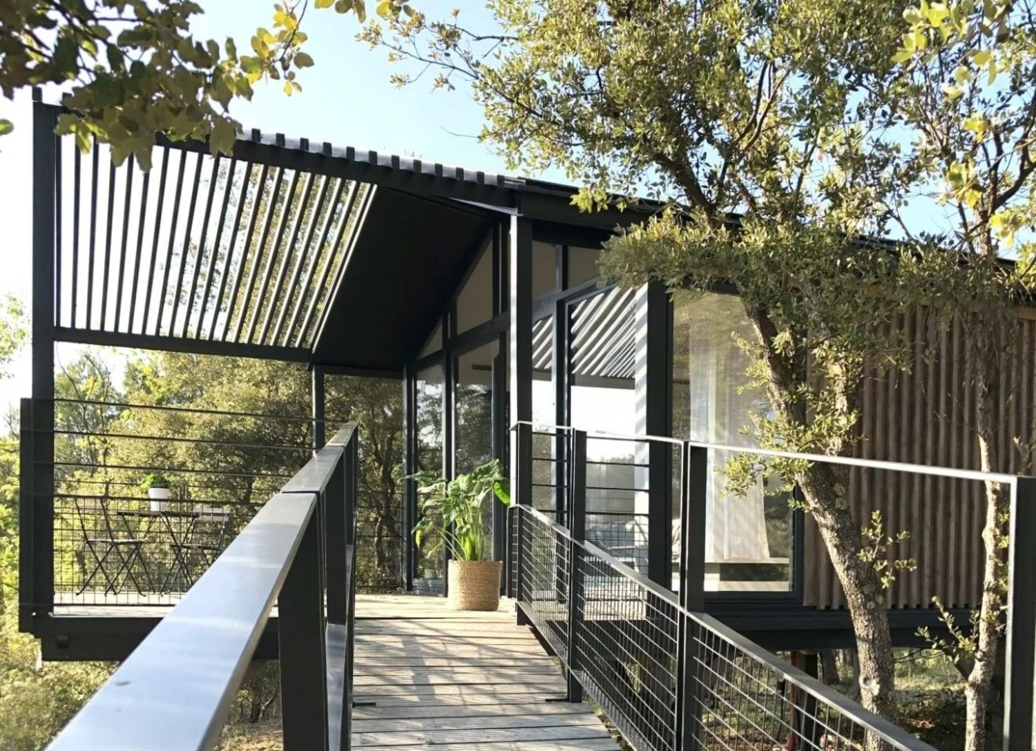 Une maison moderne en bois et en métal avec une terrasse suspendue entourée d'arbres et de plantes, sous un ciel clair.