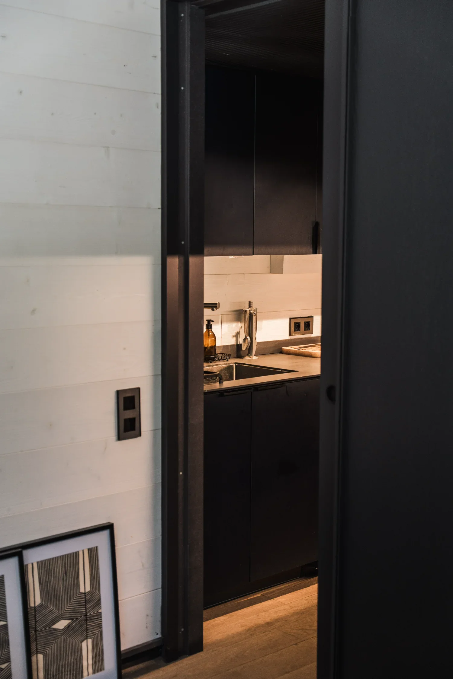 Une cuisine moderne avec des armoires noires, un évier, un robinet, une bouteille de liquide vaisselle et des prises électriques, vue à travers une porte entrouverte dans un intérieur contemporain.