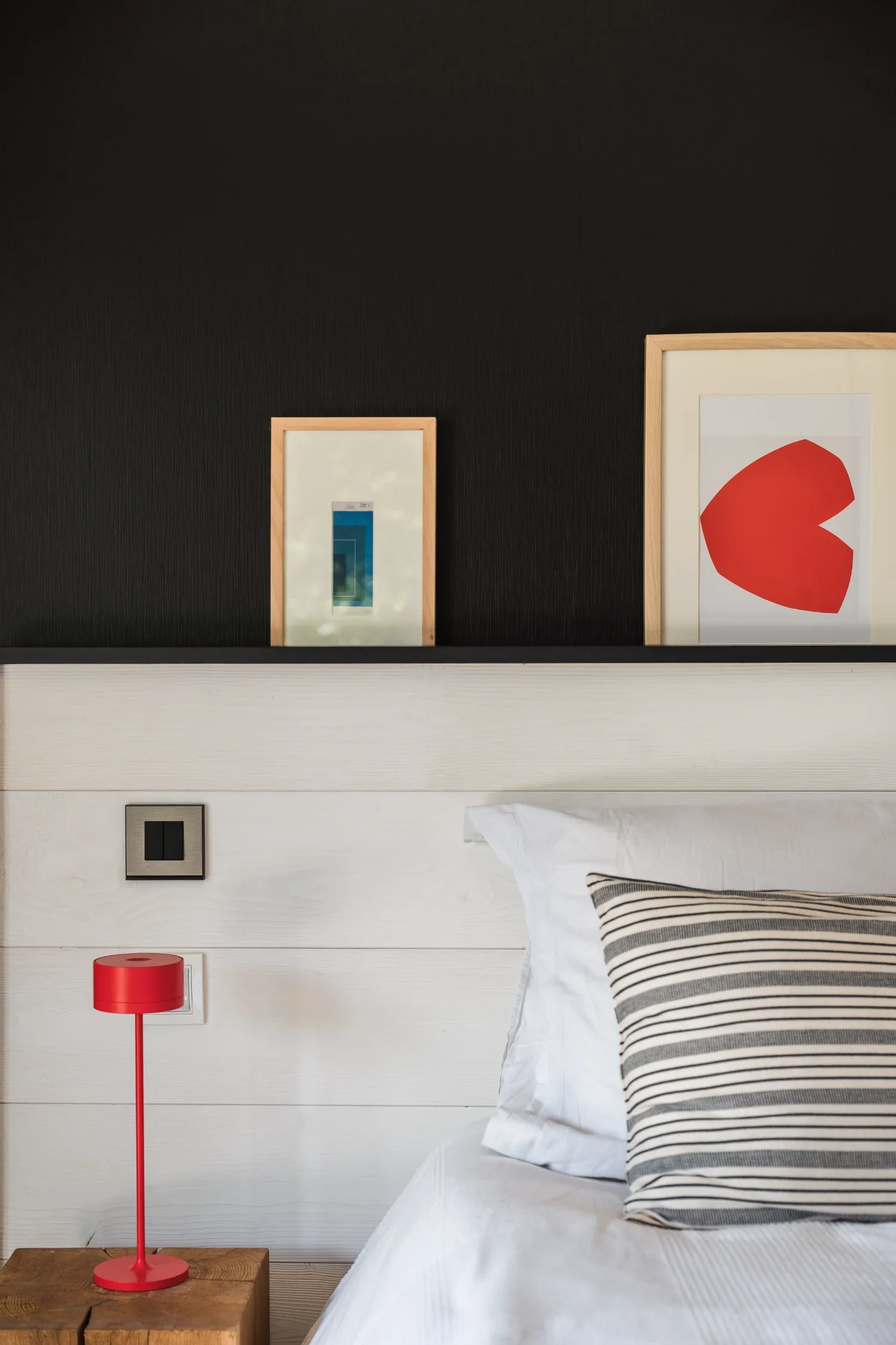 Un lit avec des oreillers blanc et à rayures dans une chambre moderne, avec des peintures encadrées sur la tête de lit, un lampadaire rouge sur une table en bois et un mur noir en arrière-plan.