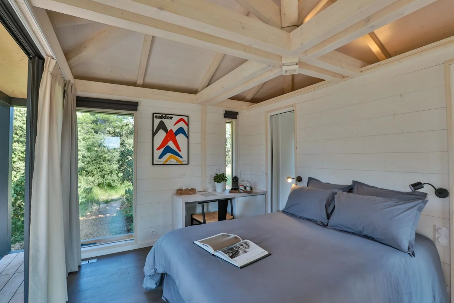 Intérieur d'un Ecolodge KOBE  avec lit , murs en bois clair, grand fenêtre donnant sur la nature, décoration minimaliste, tableau coloré sur le mur, petit bureau avec plante et machine à café.