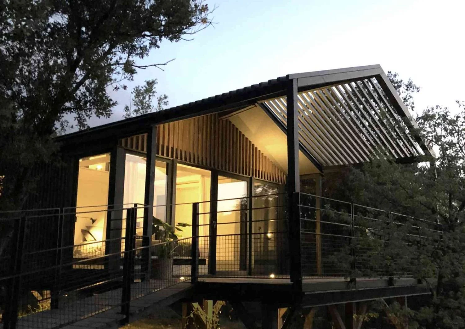 Une maison moderne en bois avec un balcon, éclairée, entourée d'arbres, la nuit.
