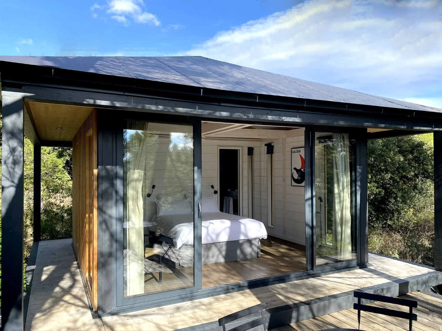Chambre avec lit blanc, murs en bois, grande fenêtre en verre, vue sur la nature, décoration moderne.