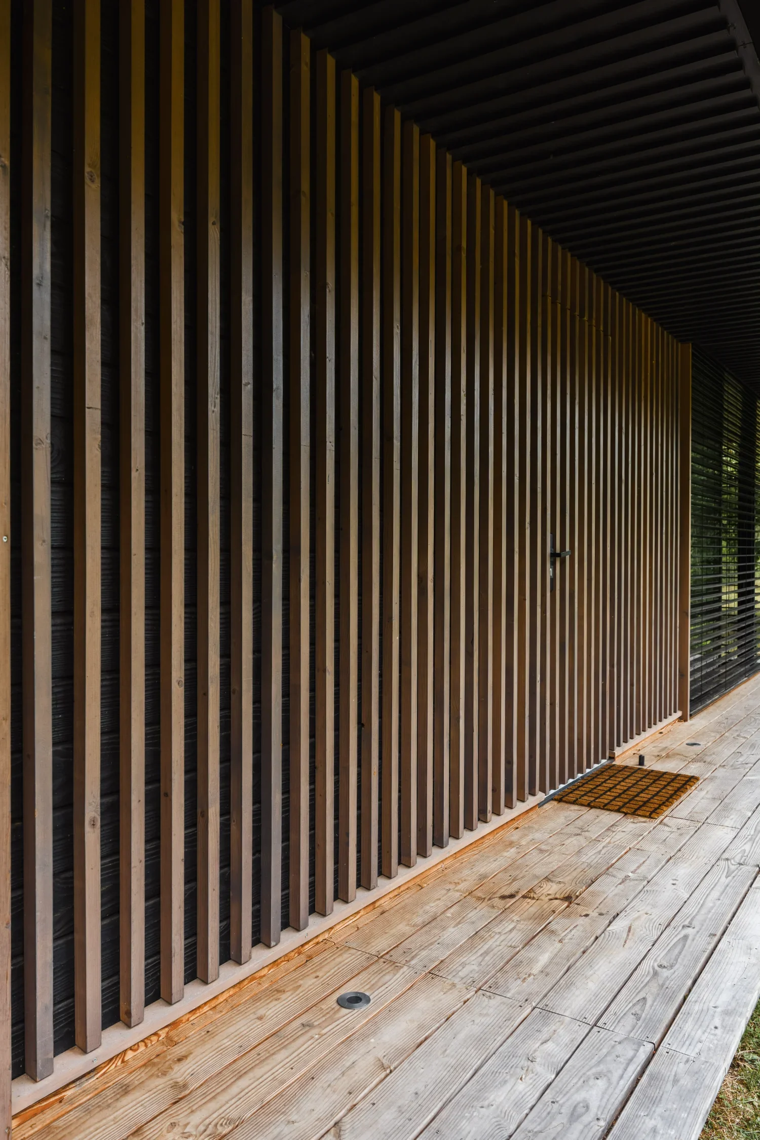Façade en bois d'un Ecolodge TESHIMA avec un mur en lattes verticales, un volet en lattes horizontales, et un paillasson en caoutchouc à l'entrée de la porte.