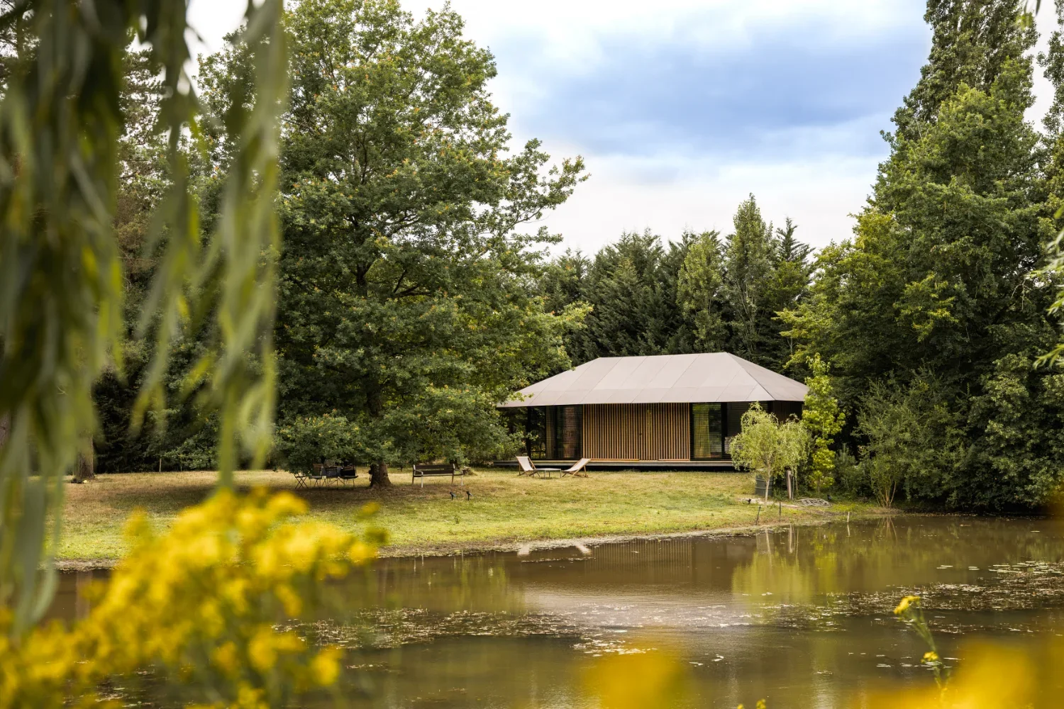 Ecolodge TESHIMA située au bord d'un plan d'eau, entourée d'arbres verts, avec un ciel nuageux en arrière-plan.