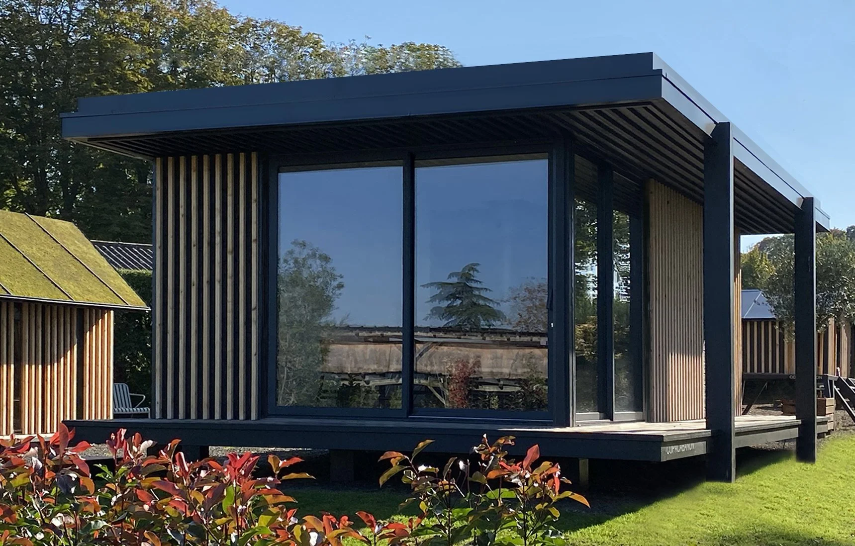 Ecolodge MEGYMA avec une façade en bois et de grandes fenêtres en verre, située sur une pelouse verte, avec des arbres en arrière-plan.