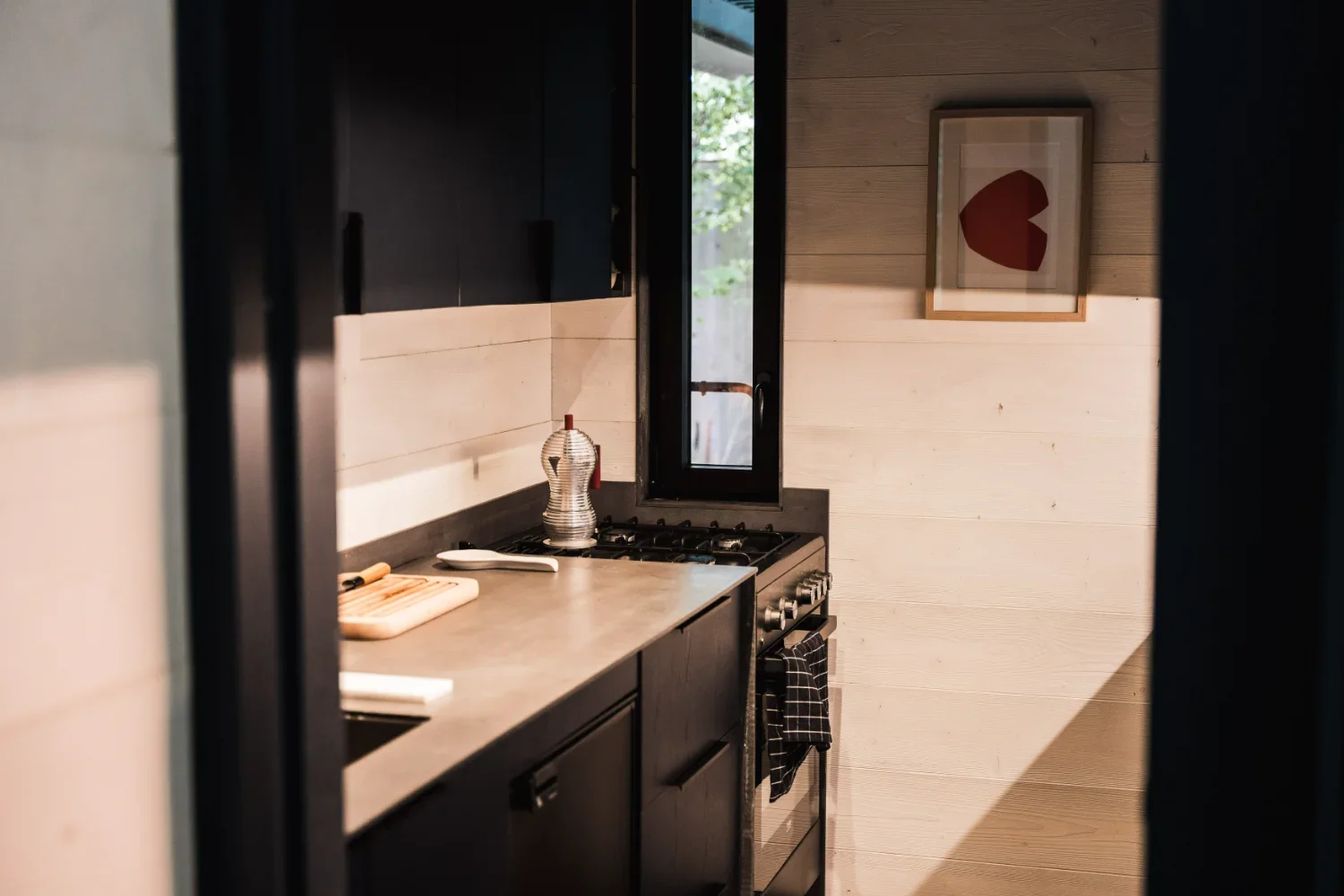 Une cuisine rustique avec un plan de travail en béton, un four à gaz noir, un mur en bois clair, une photo abstraite encadrée et une petite fenêtre.