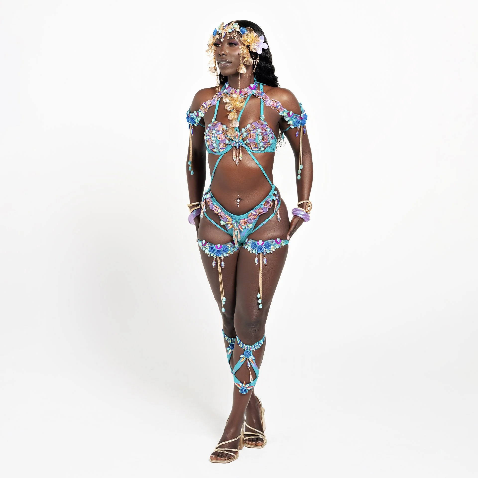 Woman wearing colorful, embellished costume with floral and jewel details and high-heeled sandals, standing against a white background.