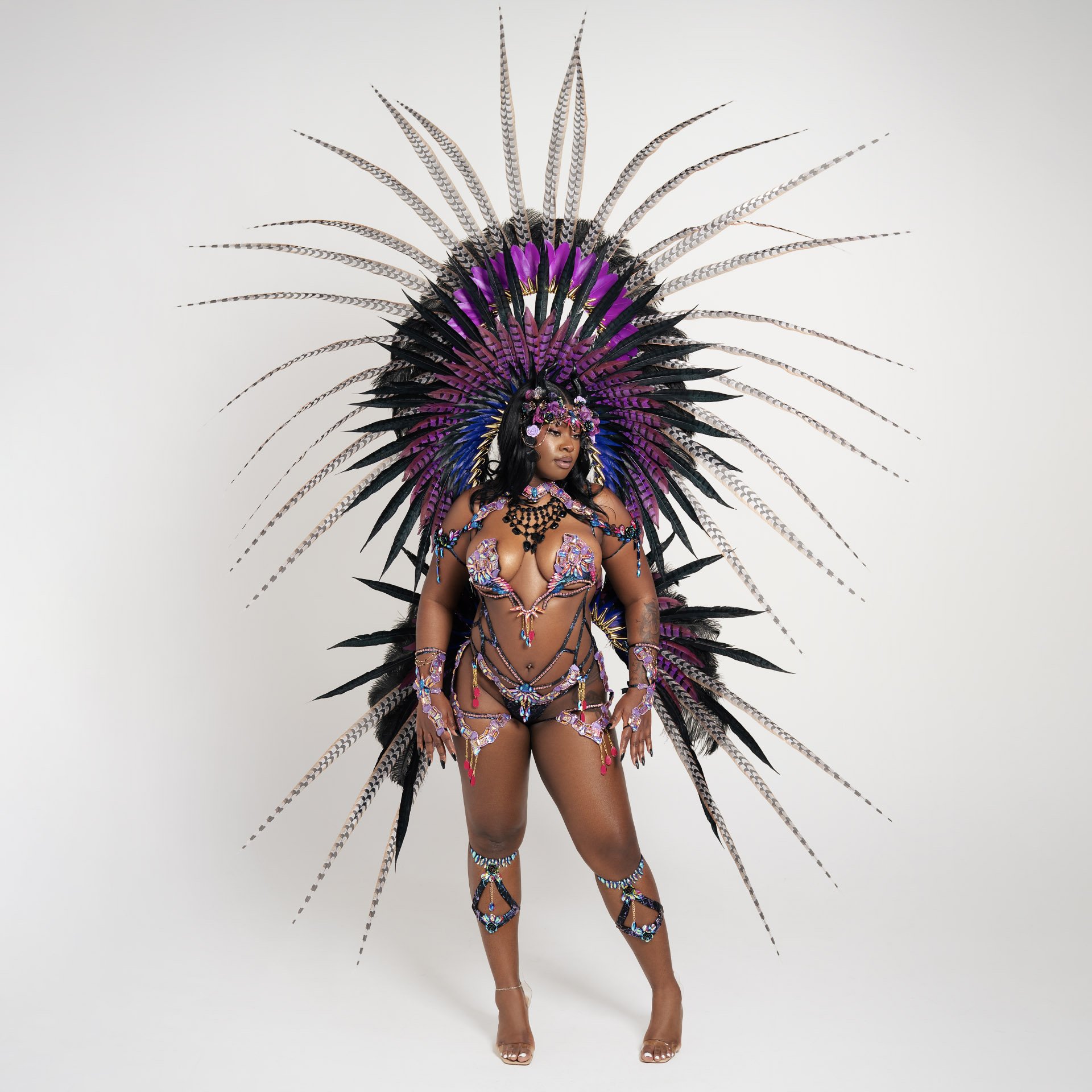A woman wearing elaborate carnival costume with purple, black, and blue feathers, beads, and jewelry, standing against a plain white background.