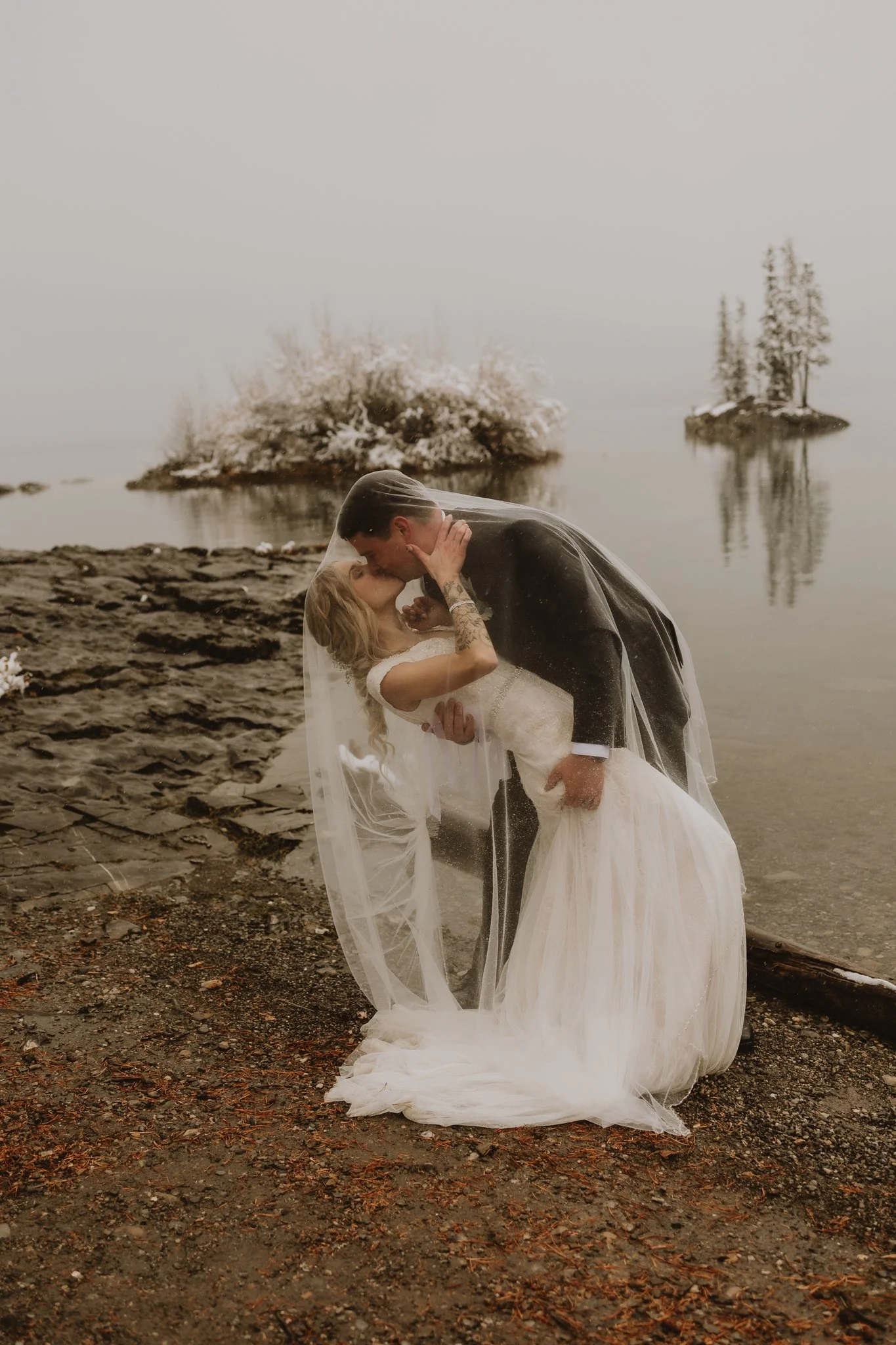 Bride and groom kissing under a veil by the water at Lake Minnewanka, Banff, Alberta, during a snowy elopement, photographed by Calgary & Alberta elopement photographer Shannon Boyd
