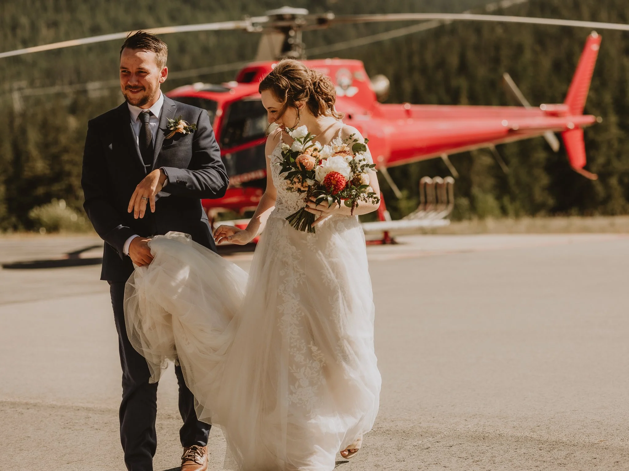 Bride and groom in wedding attire walking outside with a red helicopter in the background.