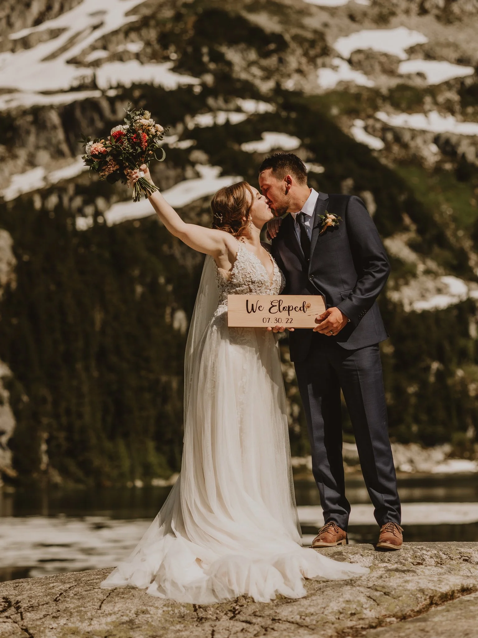 Couple celebrating on top of a mountain in Whistler, BC during their helicopter elopement, holding a ‘We Just Eloped’ sign and cheering, photographed by Alberta & BC elopement photographer Shannon Boyd