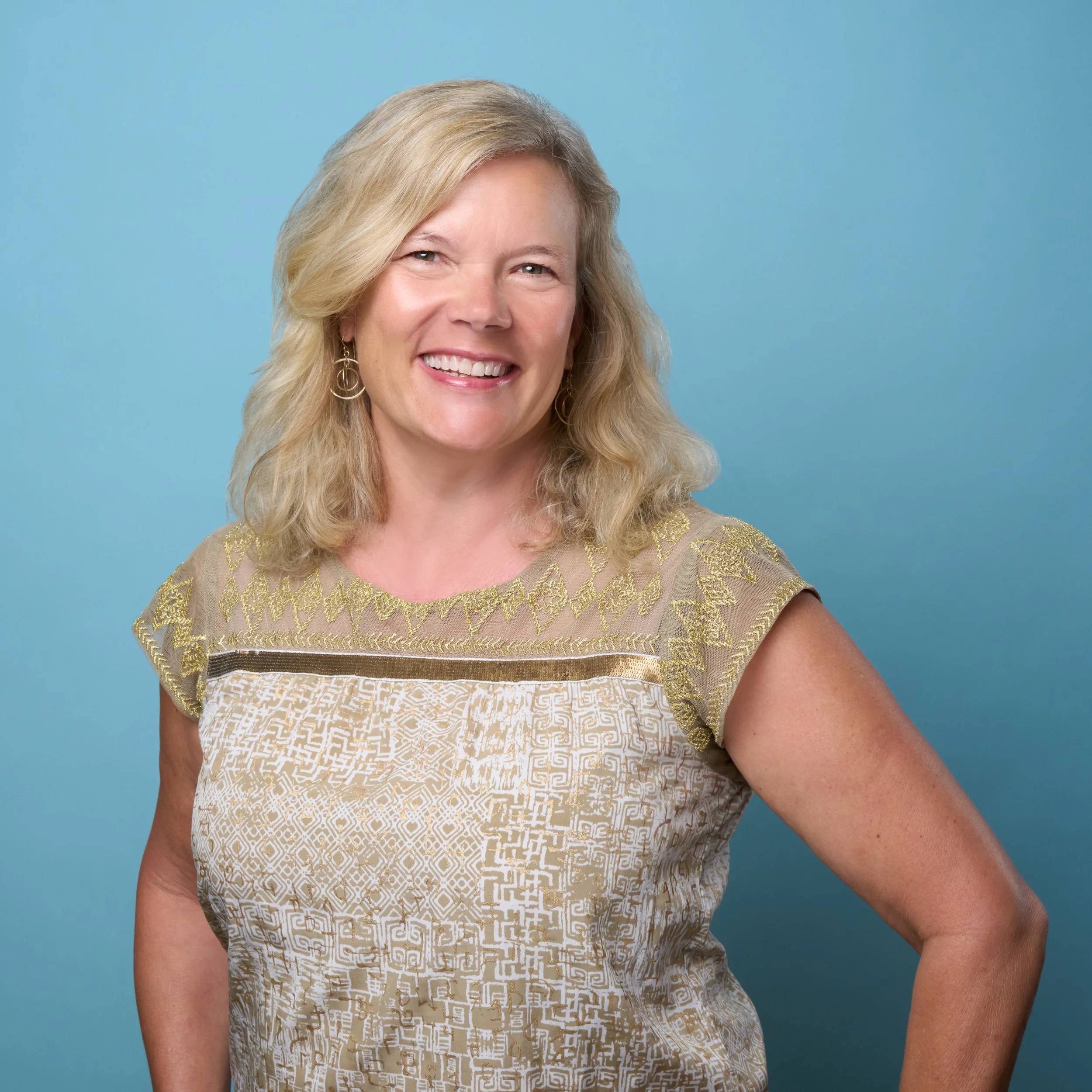 A smiling middle-aged woman with blonde hair, wearing a patterned gold and white top, standing against a light blue background.