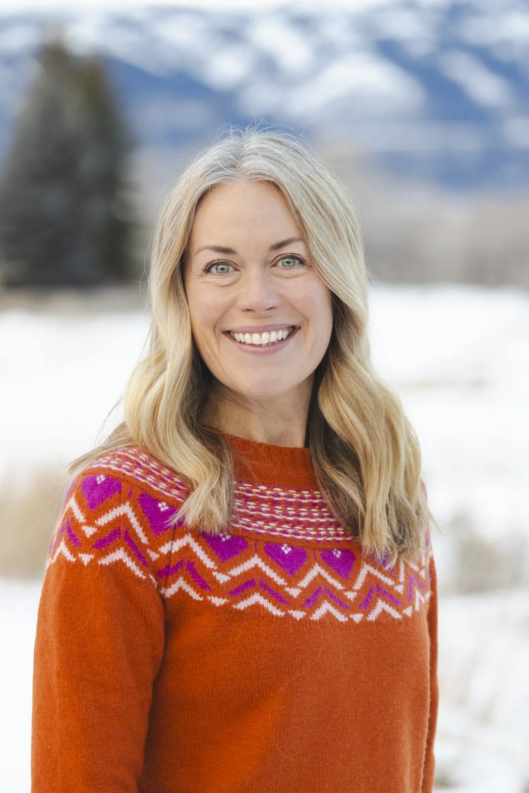 A smiling woman with blonde hair and blue eyes wearing an orange sweater with a colorful snowflake pattern outdoors in a snowy landscape.
