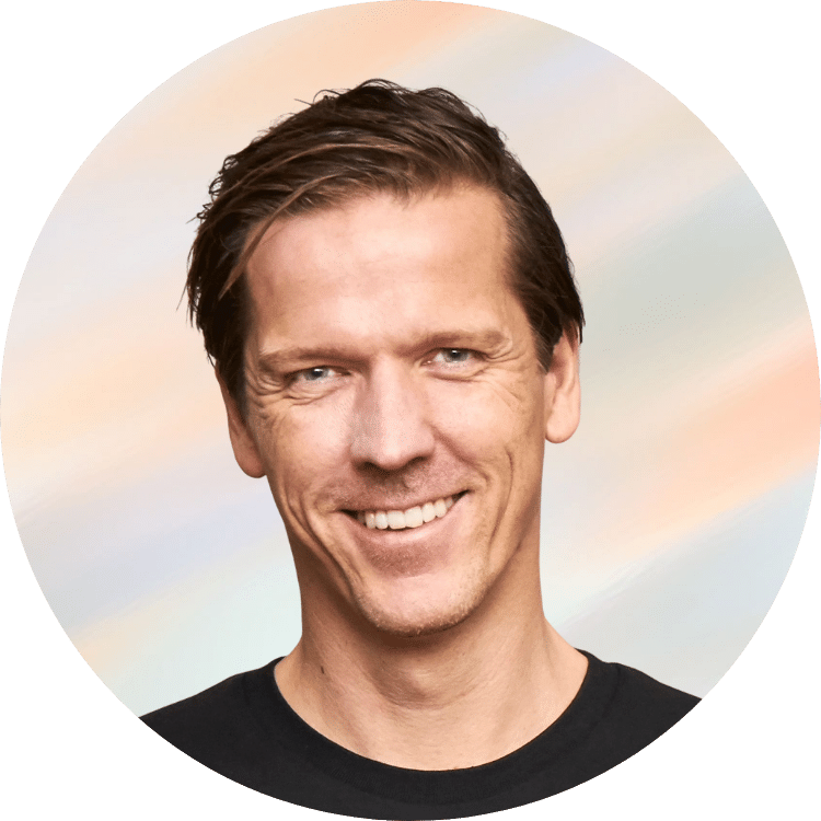 Ein Mann mit braunen Haaren und einem schwarzen T-Shirt, lächelnd, vor einem pastelfarbenen Himmel im Kreisrahmen