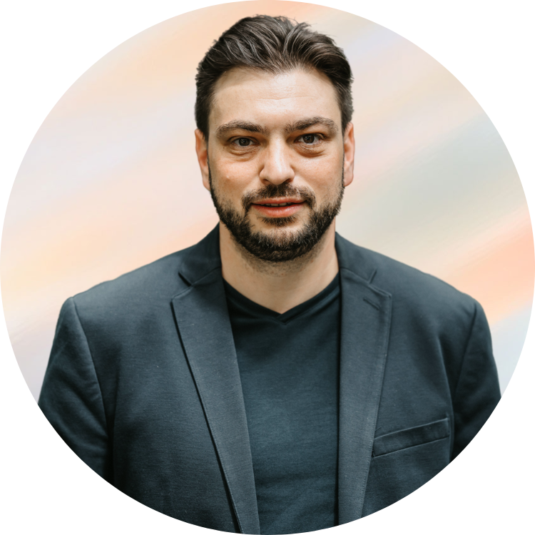 Porträt eines Mannes mit dunklem Haar, Bart und dunkler Kleidung vor einem bunten, weichen Hintergrund.