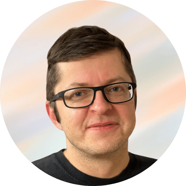 Porträt eines Mannes mit dunklem Haar, Brille und schwarzem T-Shirt vor einem pastellfarbenen Himmelshintergrund.