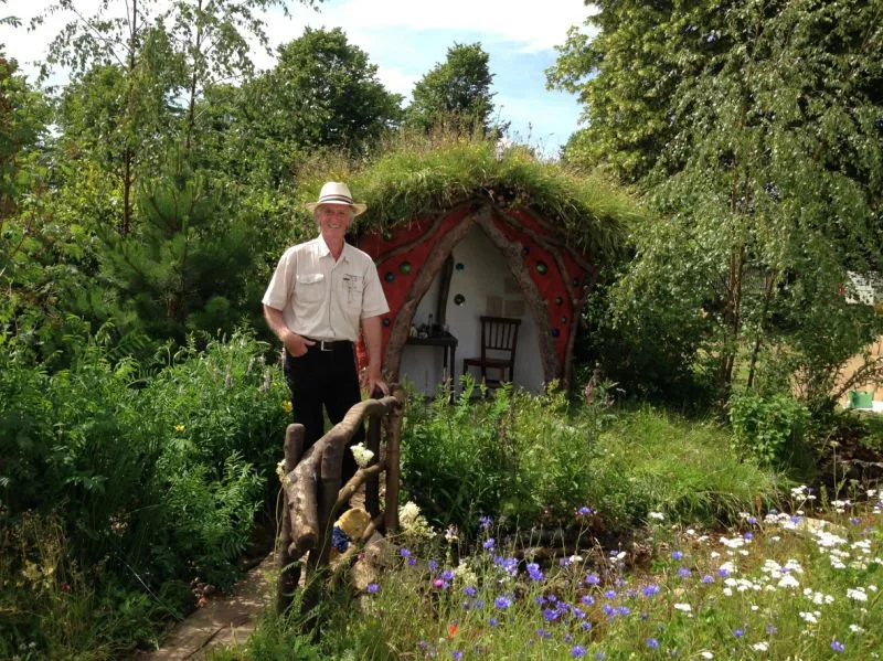 Man standing in a lush garden with a small, earth-covered, dome-shaped house decorated with colorful stones in the background.