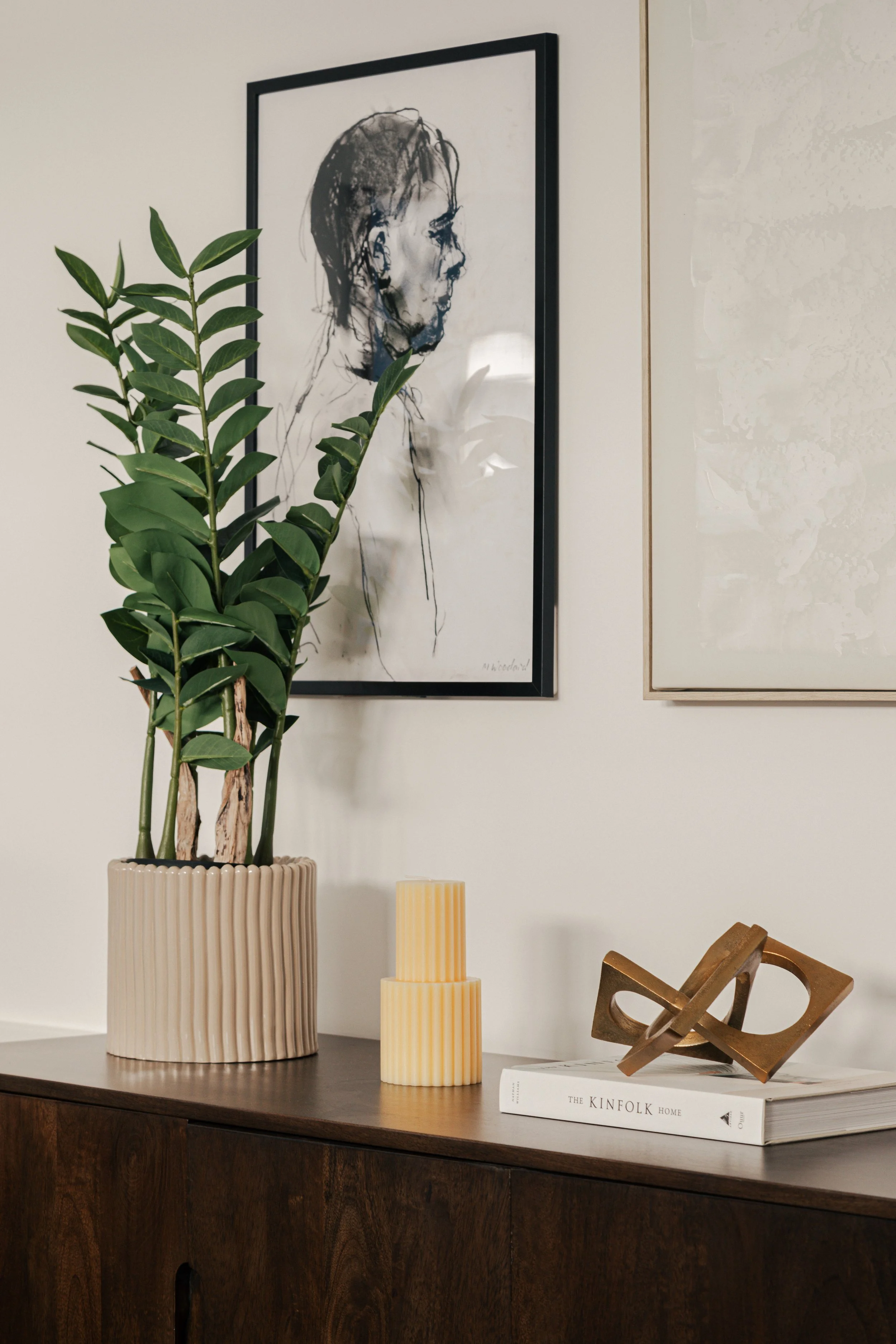 Interior decor with a framed line art portrait on the white wall, a tall green potted plant in a ribbed beige planter, two yellow ribbed candles, a decorative wooden sculpture, and a large book titled 'The Kinfolk Home' on a dark wooden sideboard.