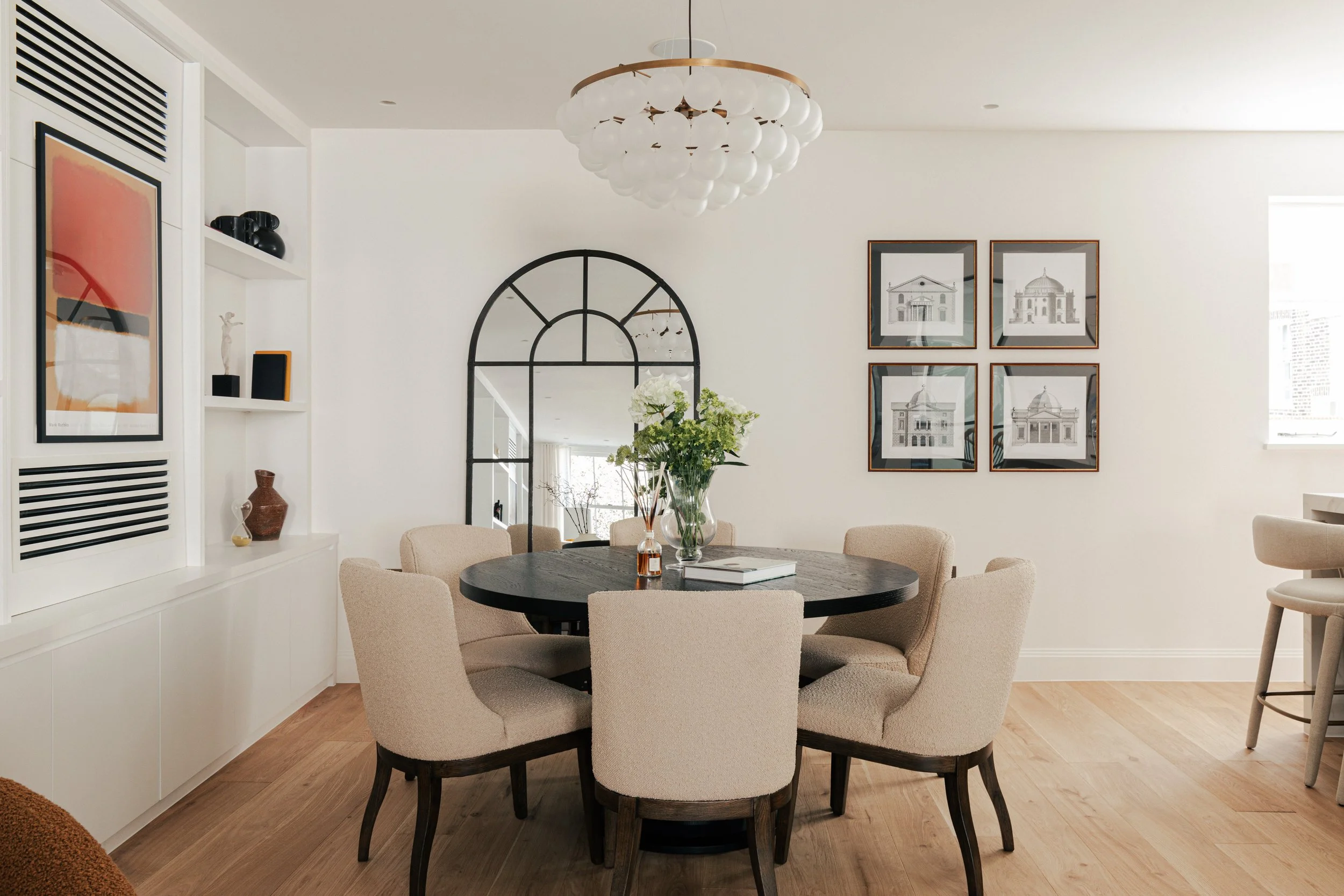 Modern dining room with a round black table surrounded by beige upholstered chairs, a vase of white and green flowers, bookshelf with artwork and decorative items, black metal arch mirror, framed architectural prints on the wall, light wood flooring,