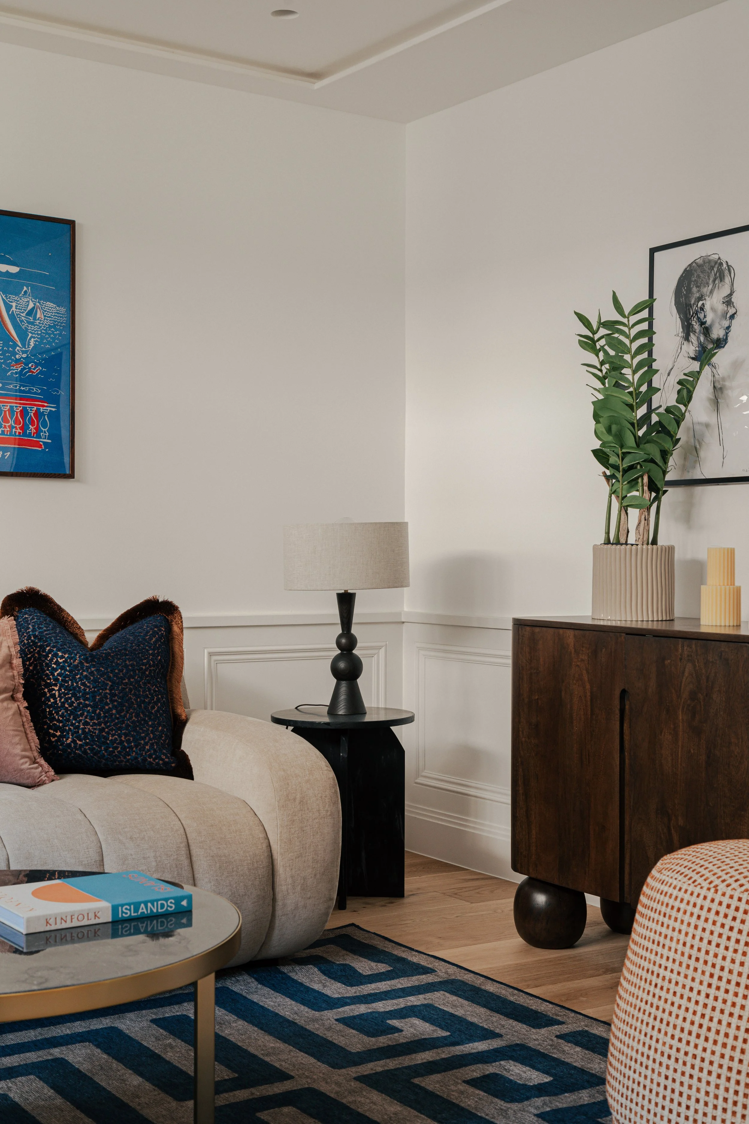 Living room corner with beige sofa, black side table with a lamp, wooden cabinet with a plant, framed artwork on the wall, and a blue patterned rug.