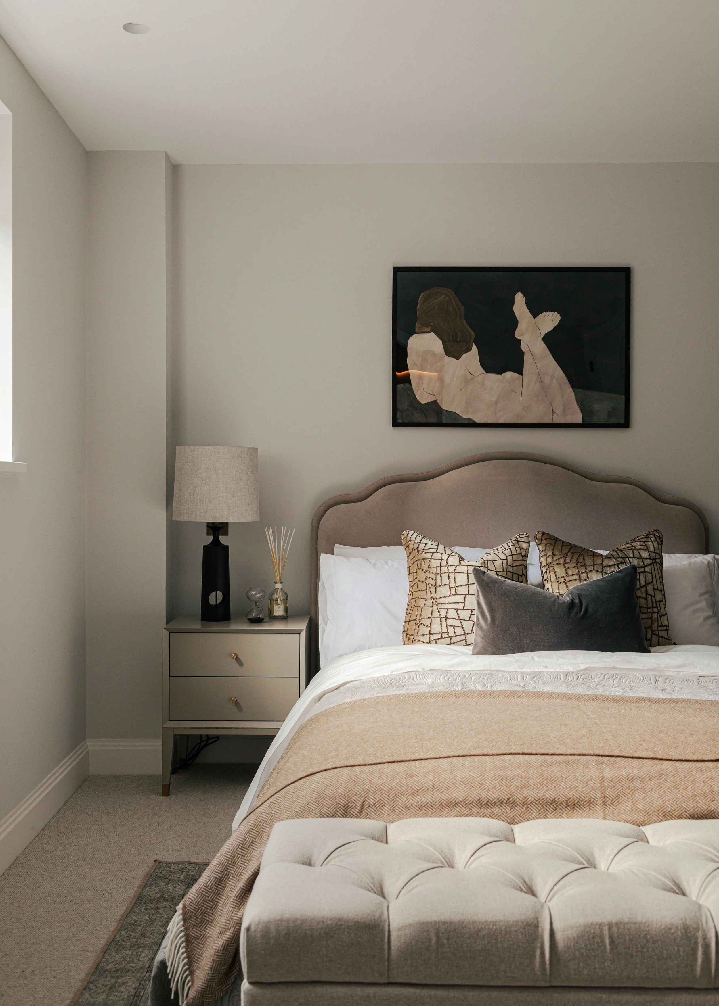 A neatly made bedroom with beige and white bedding, patterned pillows, a beige headboard, a nightstand with a lamp and decorative items, and a painting of a reclining person above the bed.