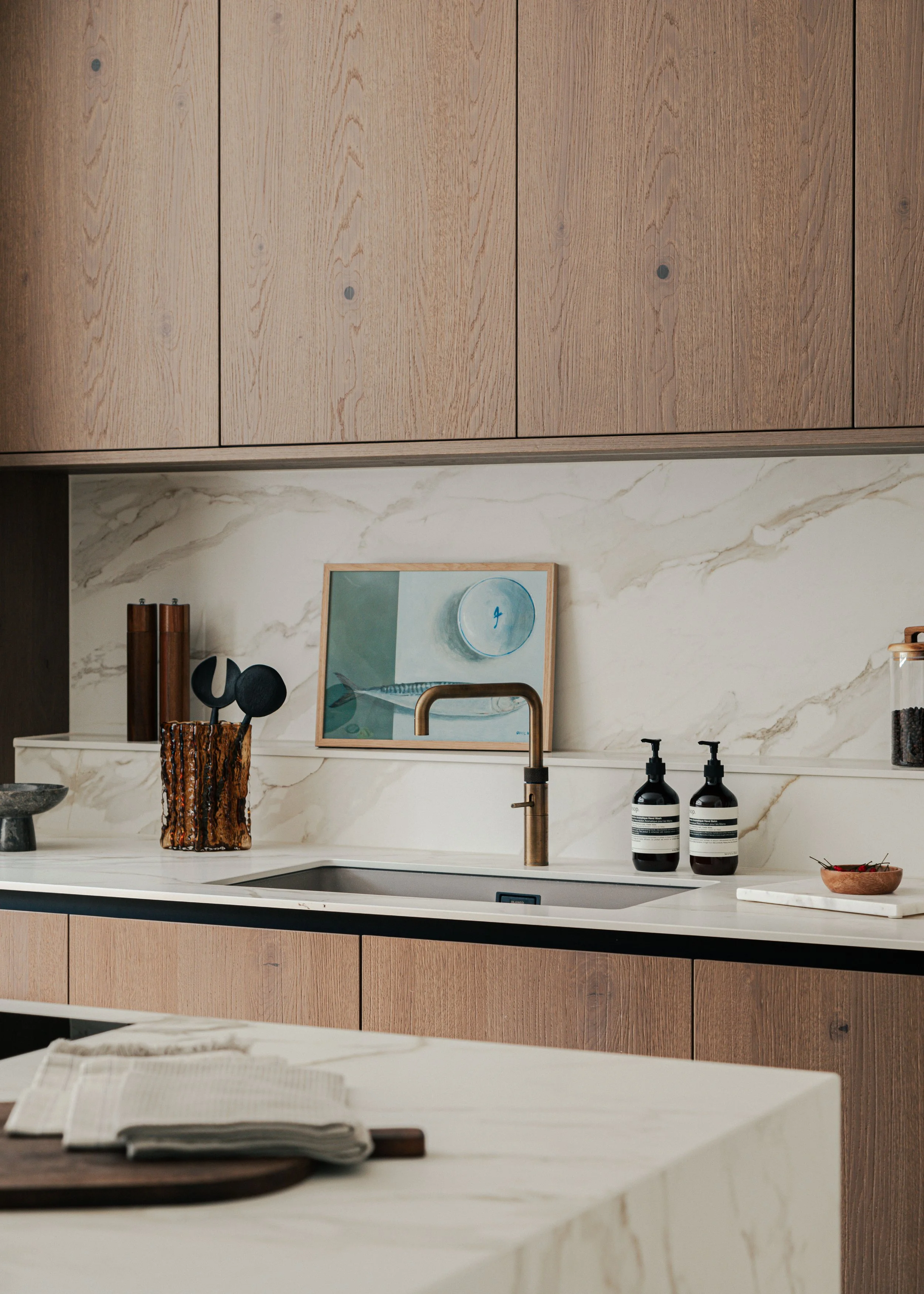 Modern kitchen with wooden cabinets, marble backsplash, and a white countertop, featuring a framed artwork, soap dispensers, and kitchen utensils.