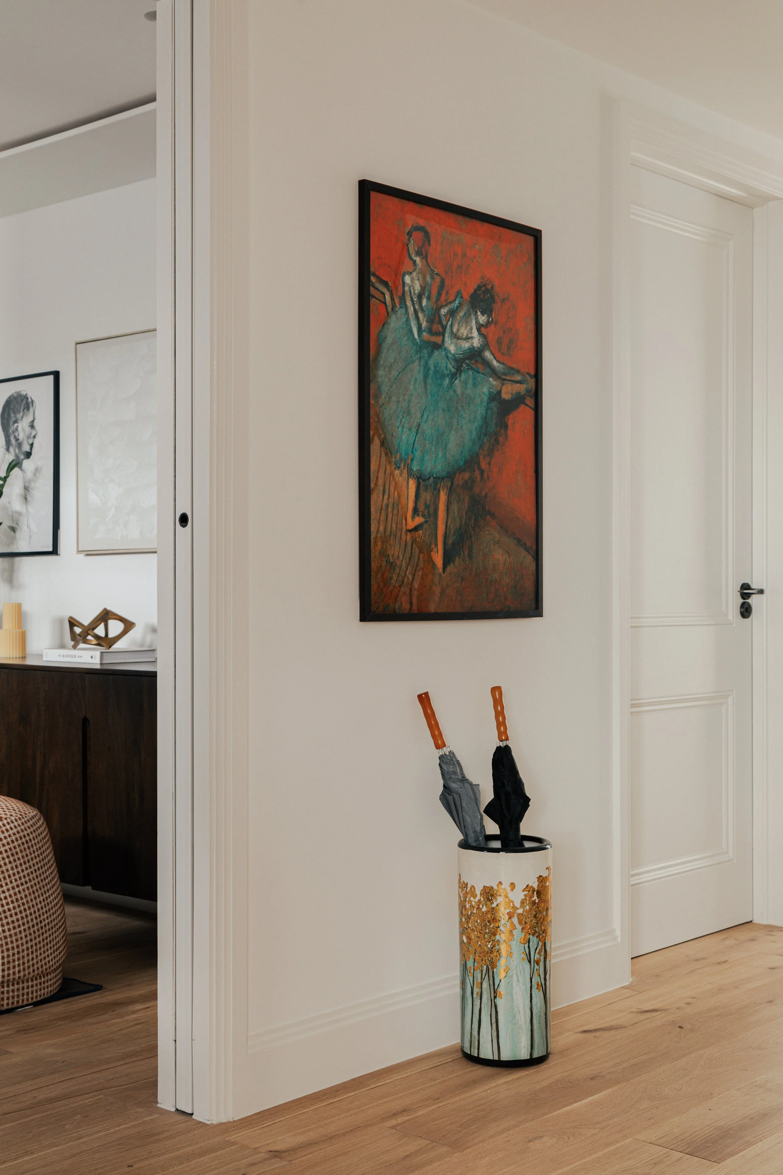 Interior wall with a framed painting of two ballerinas and a decorative umbrella stand holding two umbrellas.