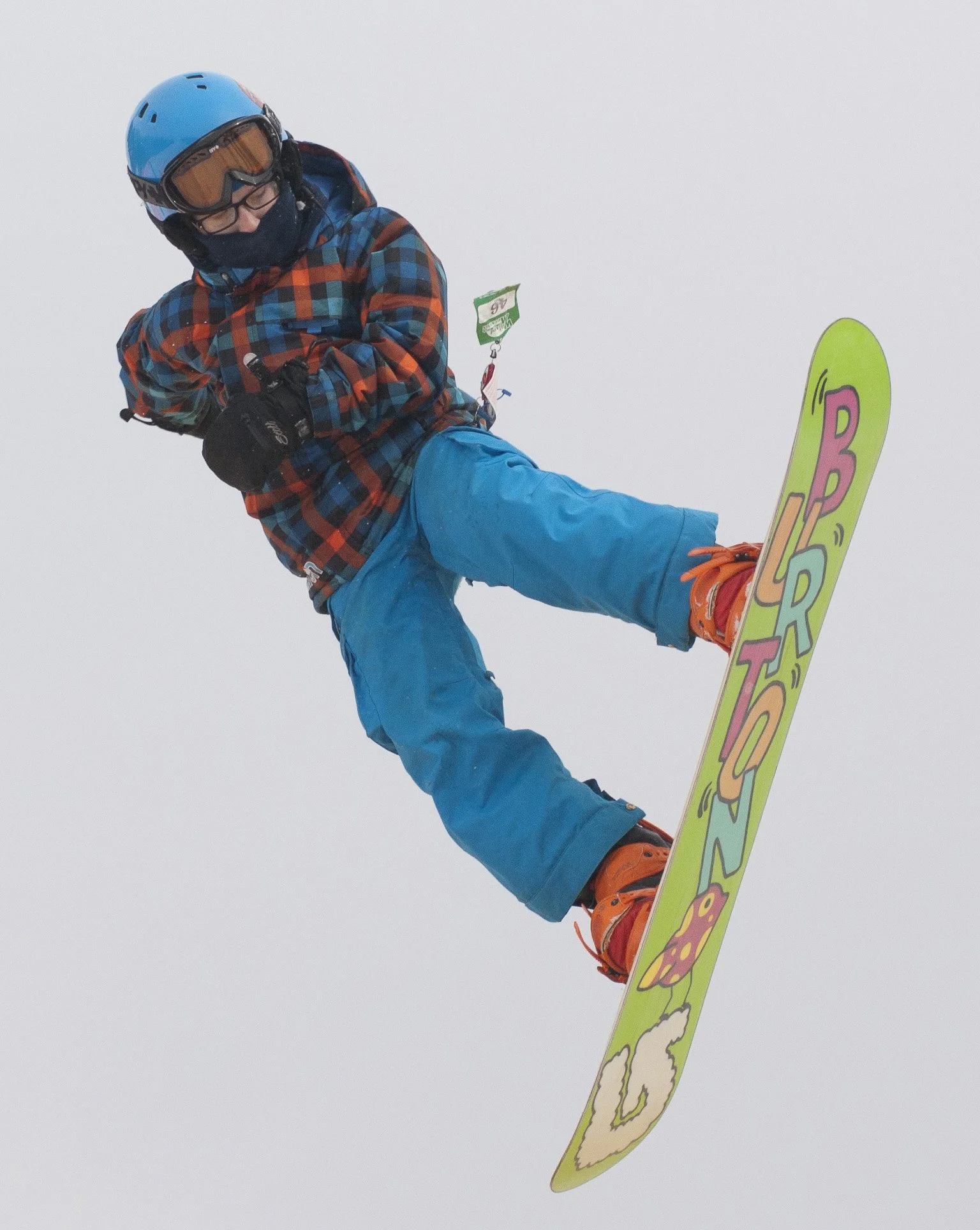 Snowboarder wearing a blue helmet, colorful checkered jacket, blue pants, and orange boots performing a mid-air jump against a white sky.
