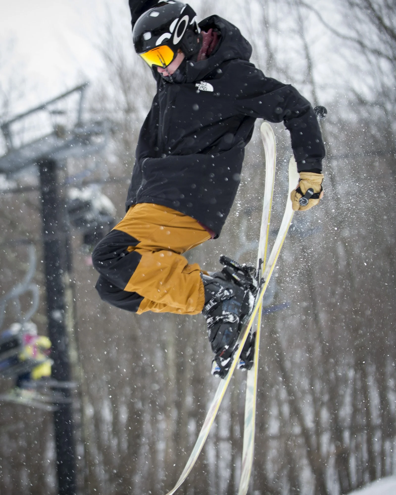 A person wearing a black helmet, black jacket, and orange snow pants is performing a jump on skis in a snowy outdoor setting.